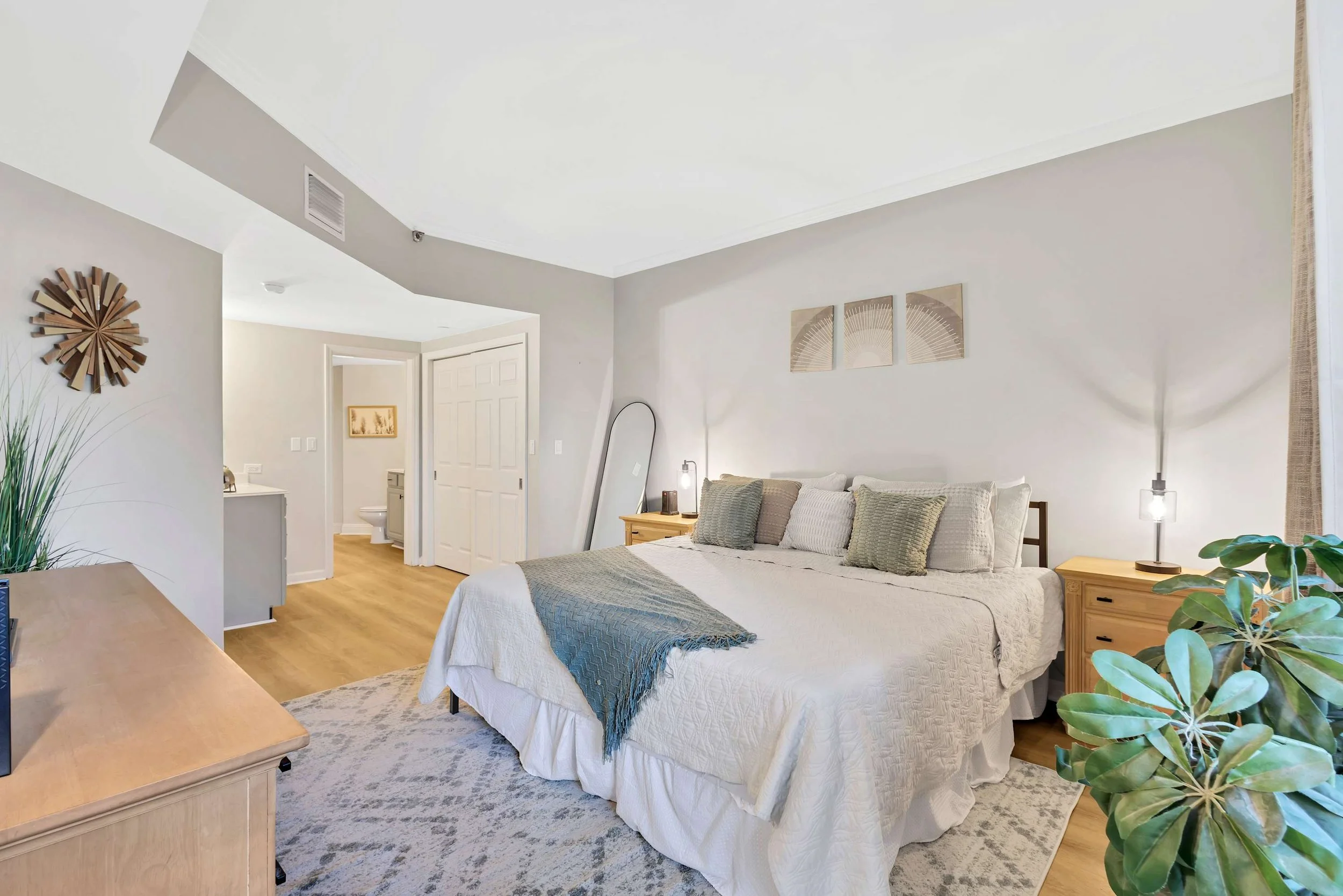 Modern bedroom with a king-size bed, two nightstands, and a decorative wall piece. The room features a neutral color palette with a plant in the corner, a full-length mirror, and recessed lighting. The bathroom is visible through an open doorway.