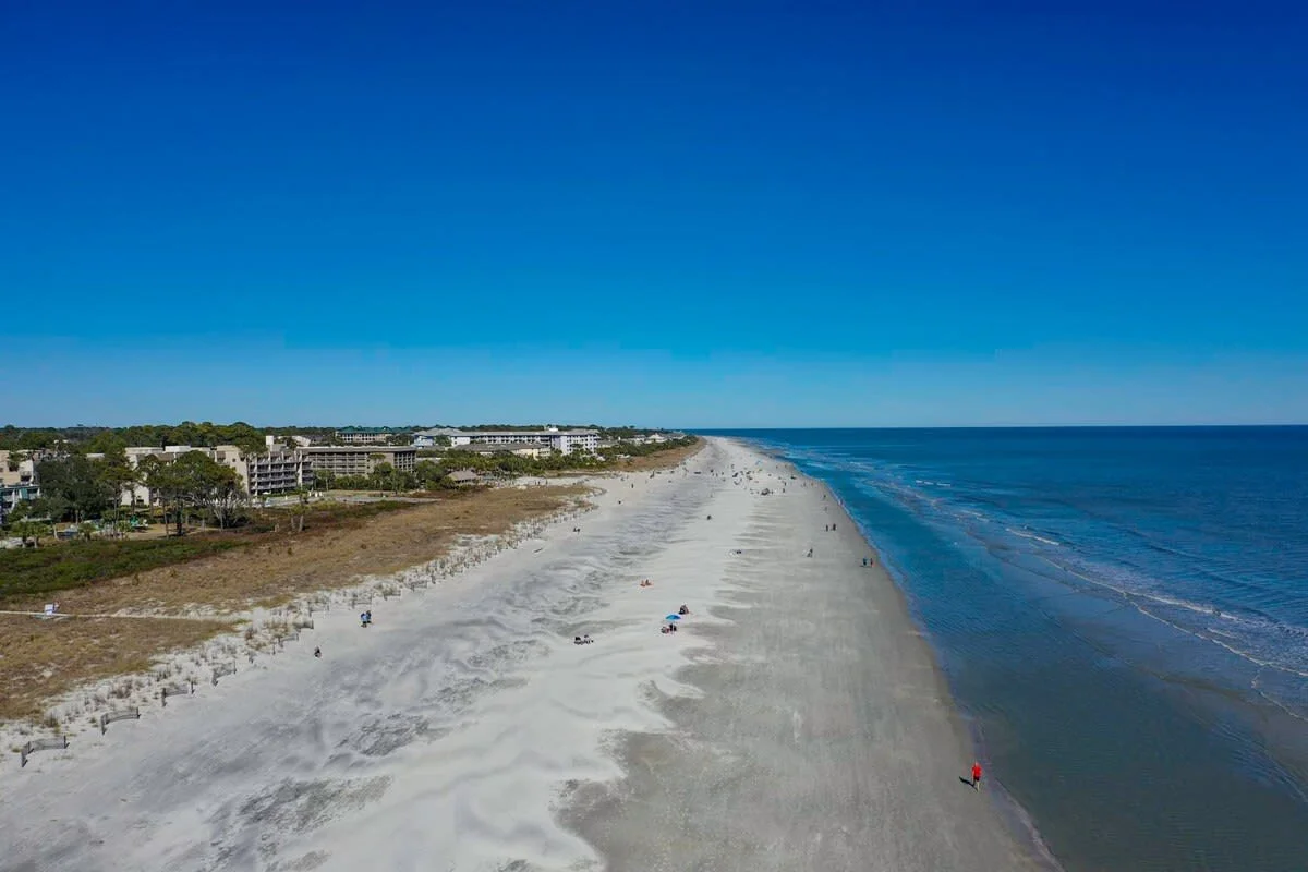 Aerial view of a sandy beach with people, ocean, and distant buildings under a clear blue sky.