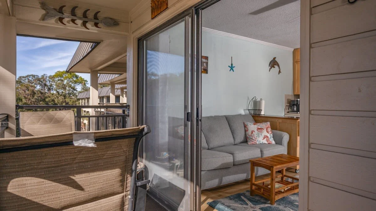 Interior of a cozy living room with a gray sofa, decorative pillows, wooden coffee table, blue starfish wall decor, and a dolphin wall sculpture. The room has a sliding glass door leading to a balcony with outdoor seating. Trees and a blue sky are vi