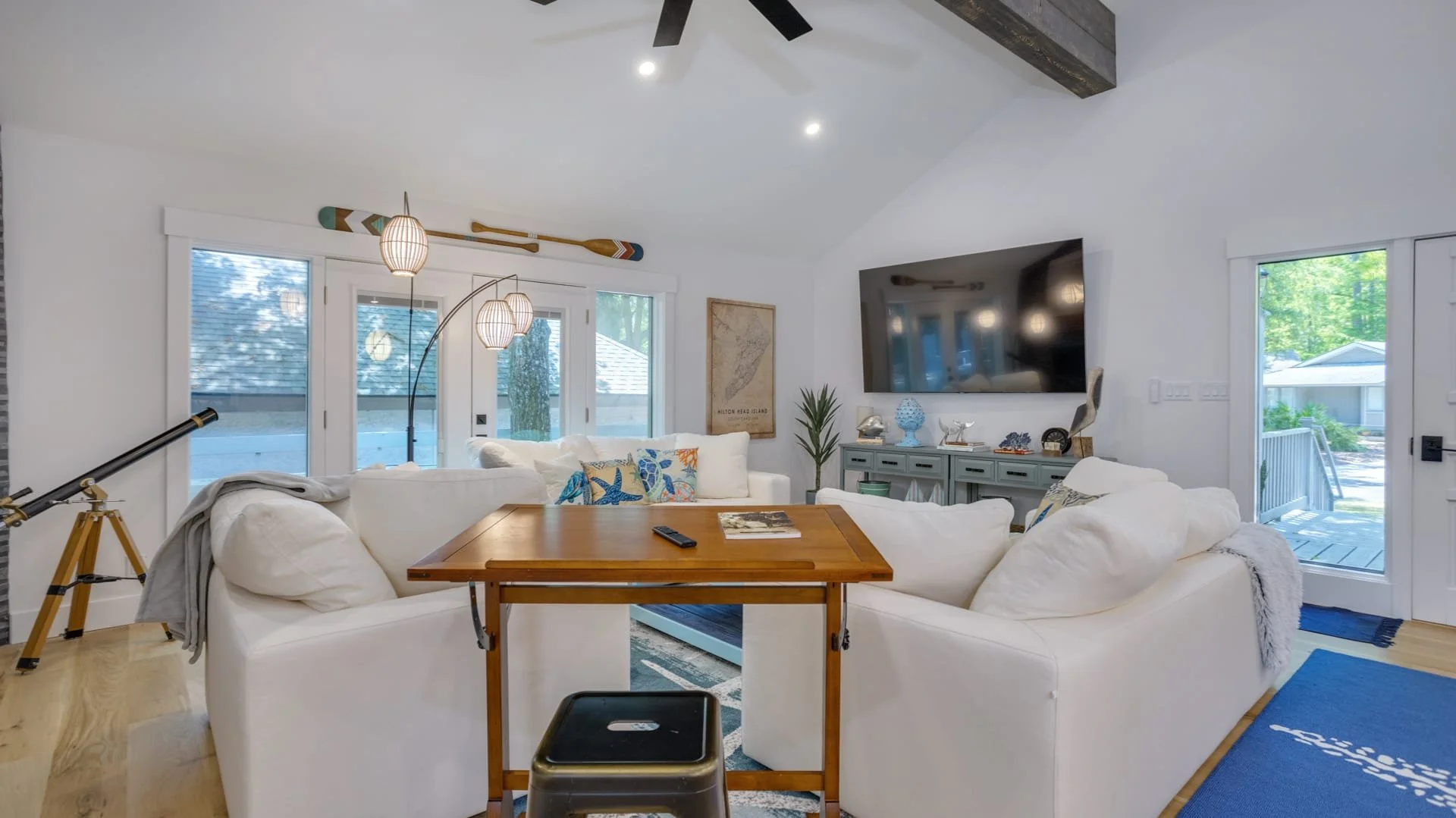 Modern living room with white sofas, a large TV, decorative map, and a telescope by the window. A wooden table with a stool and pendant lights are also present, along with nautical decor and a plant.