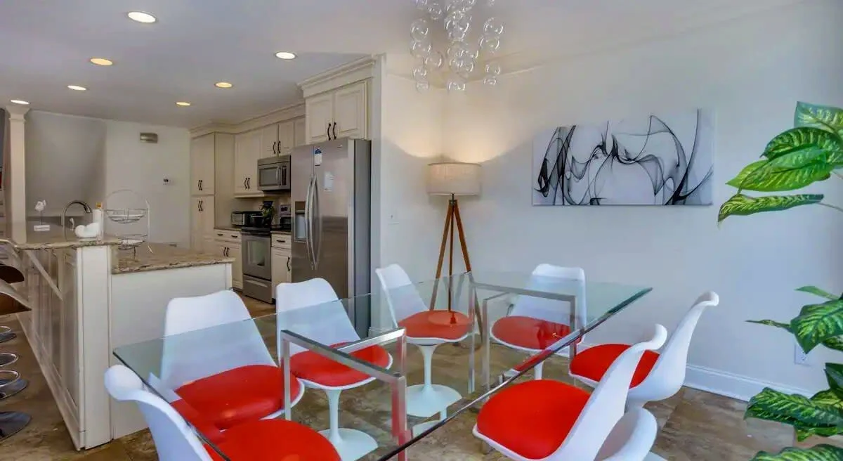 Modern kitchen and dining area with glass table, red and white chairs, stainless steel appliances, and abstract artwork on the wall.