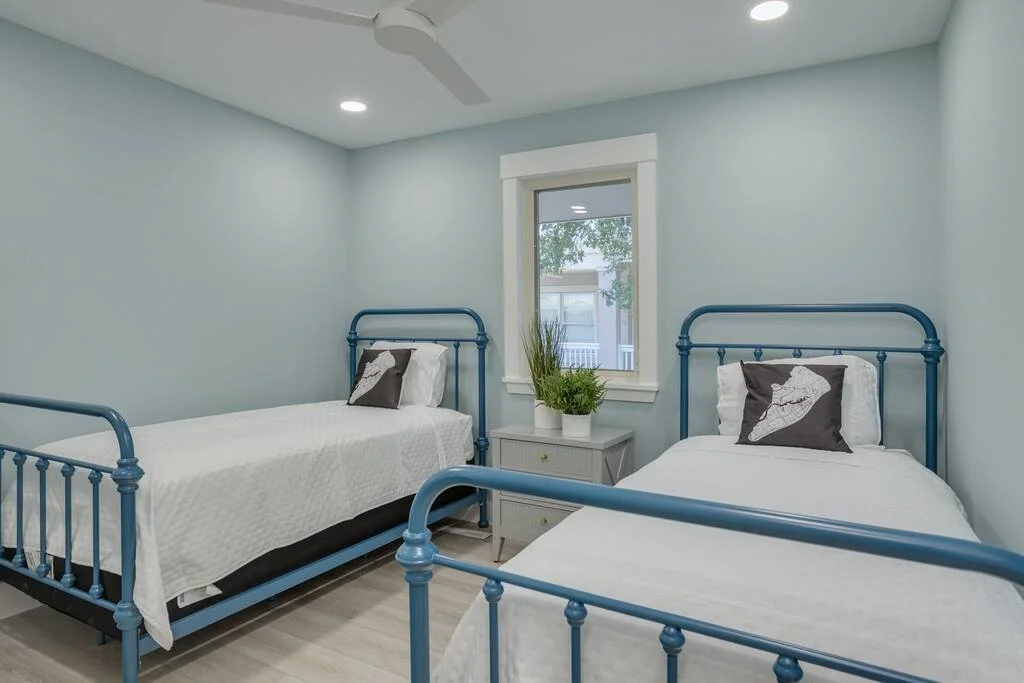 Bedroom with two twin beds featuring blue metal frames, white bedding, and decorative pillows. A small nightstand with green plants is between the beds. The walls are painted light blue, and there's a window with a view of outside greenery.