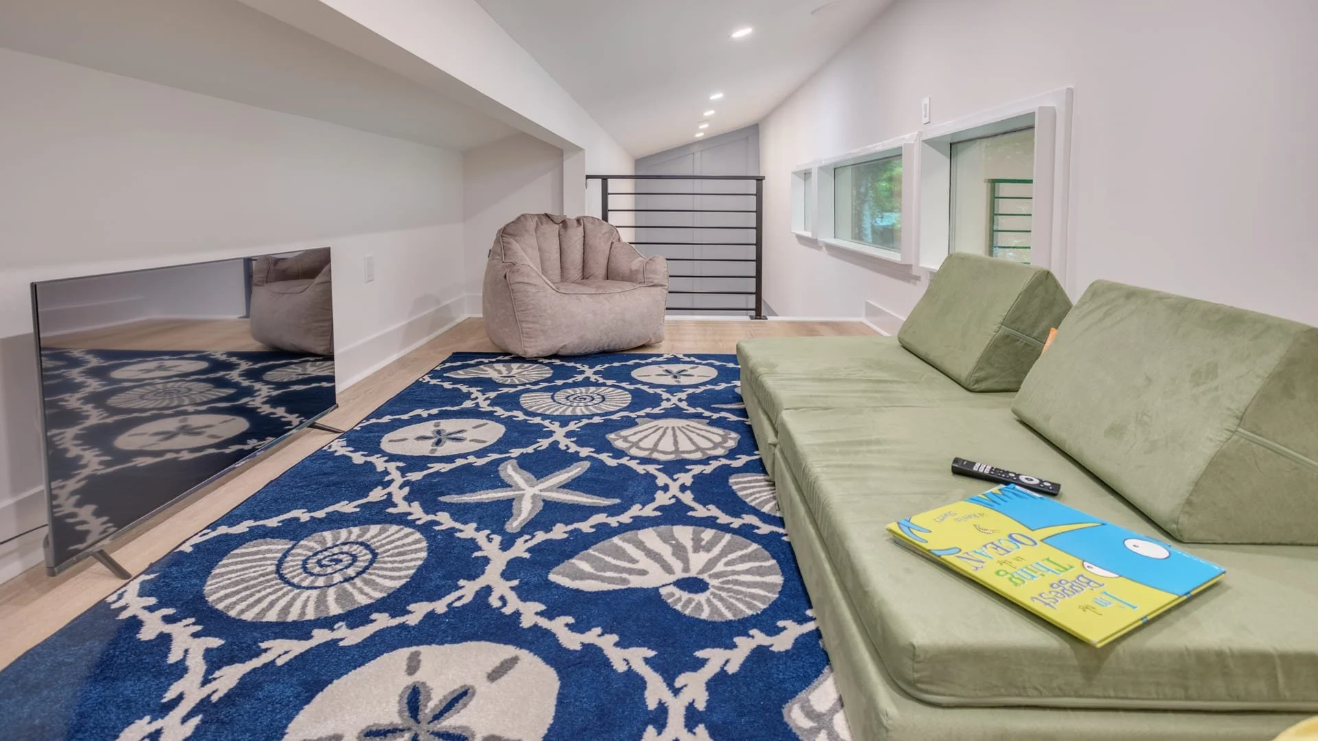 Modern loft room with green couch, blue patterned rug, beanbag chair, flat-screen TV, and windows.
