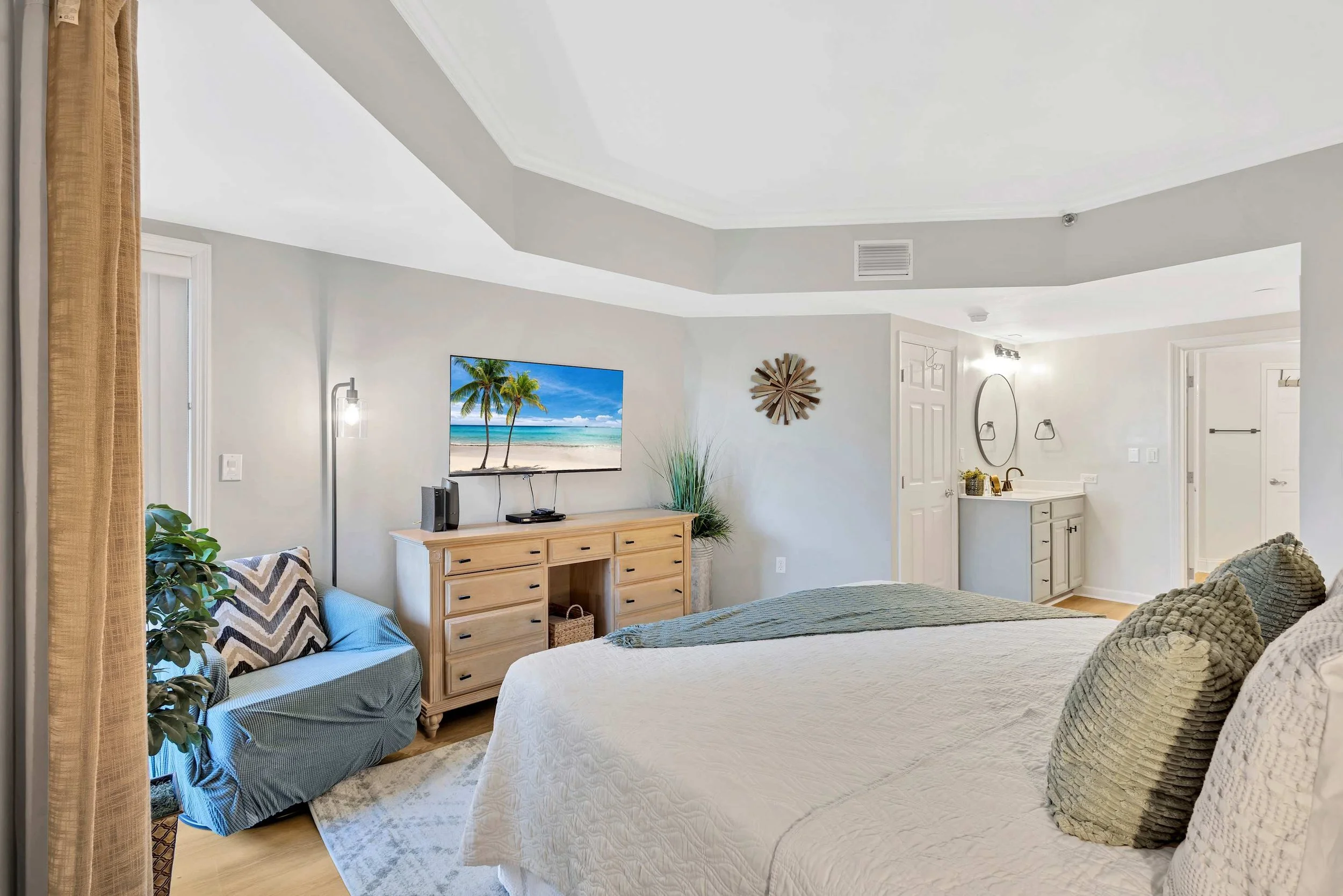Bright bedroom with a bed, decorative pillows, and dresser with a TV displaying a beach scene. A small seating area with a plant is next to the dresser. A wall-mounted art piece and a doorway leading to a bathroom with a vanity and mirror are visible