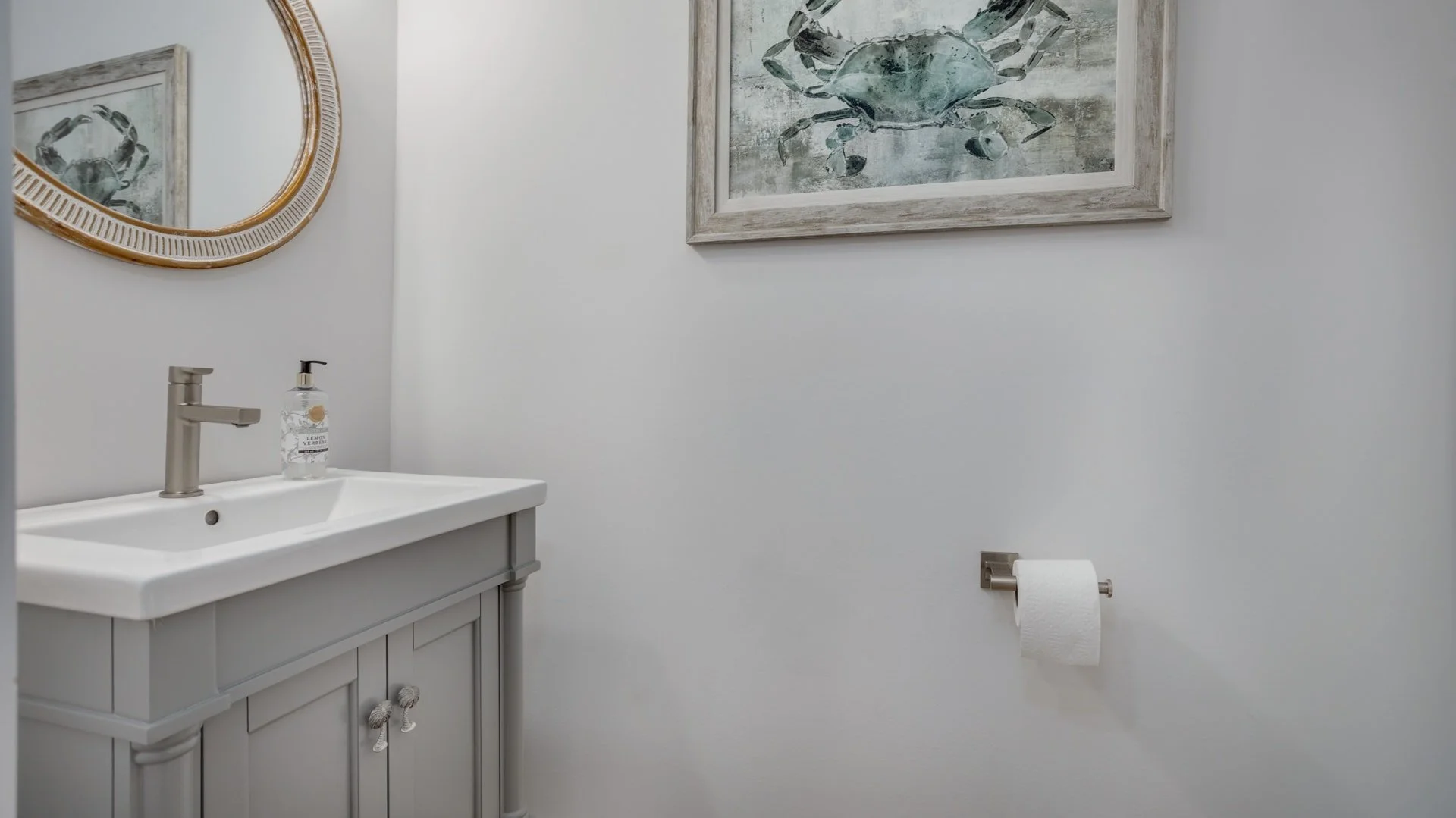 Bathroom with a gray vanity, white sink, modern faucet, soap dispenser, round mirror, framed artwork of a crab, and toilet paper holder.