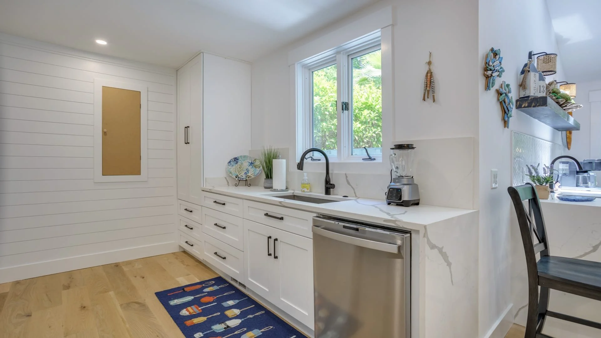 Modern kitchen with white cabinets, stainless steel appliances, and wooden flooring. Features include a countertop with a blender, a decorative plate, a window view, and wall decor. A blue rug with colorful patterns is on the floor.