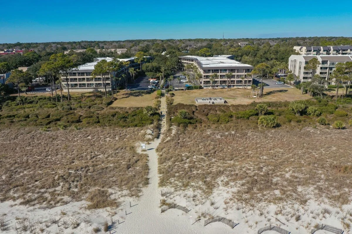 Aerial view of beachfront resort with sand dunes, a walkway leading to buildings, and surrounding greenery under clear blue skies.