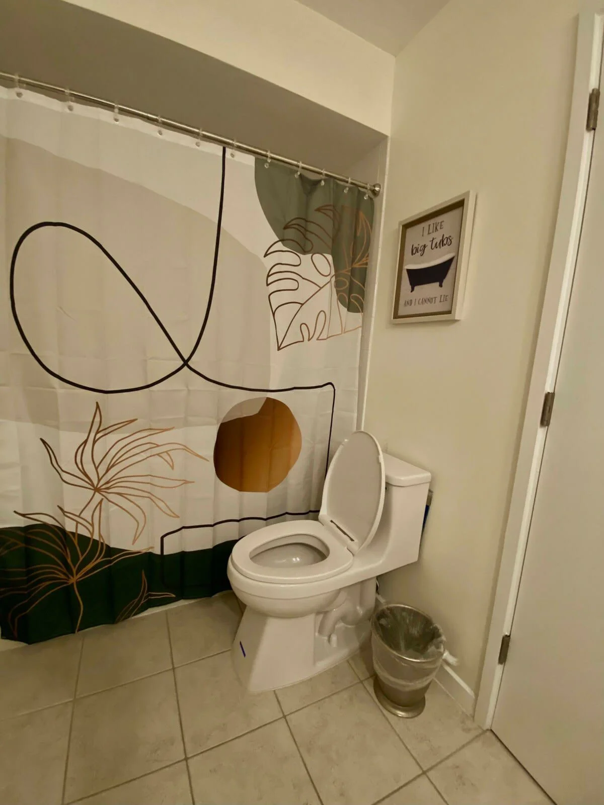 Bathroom with toilet, decorative shower curtain, and framed wall art. The curtain features abstract plant designs, and the wall art says "I like big tubs and I cannot lie." A small trash can is beside the toilet.