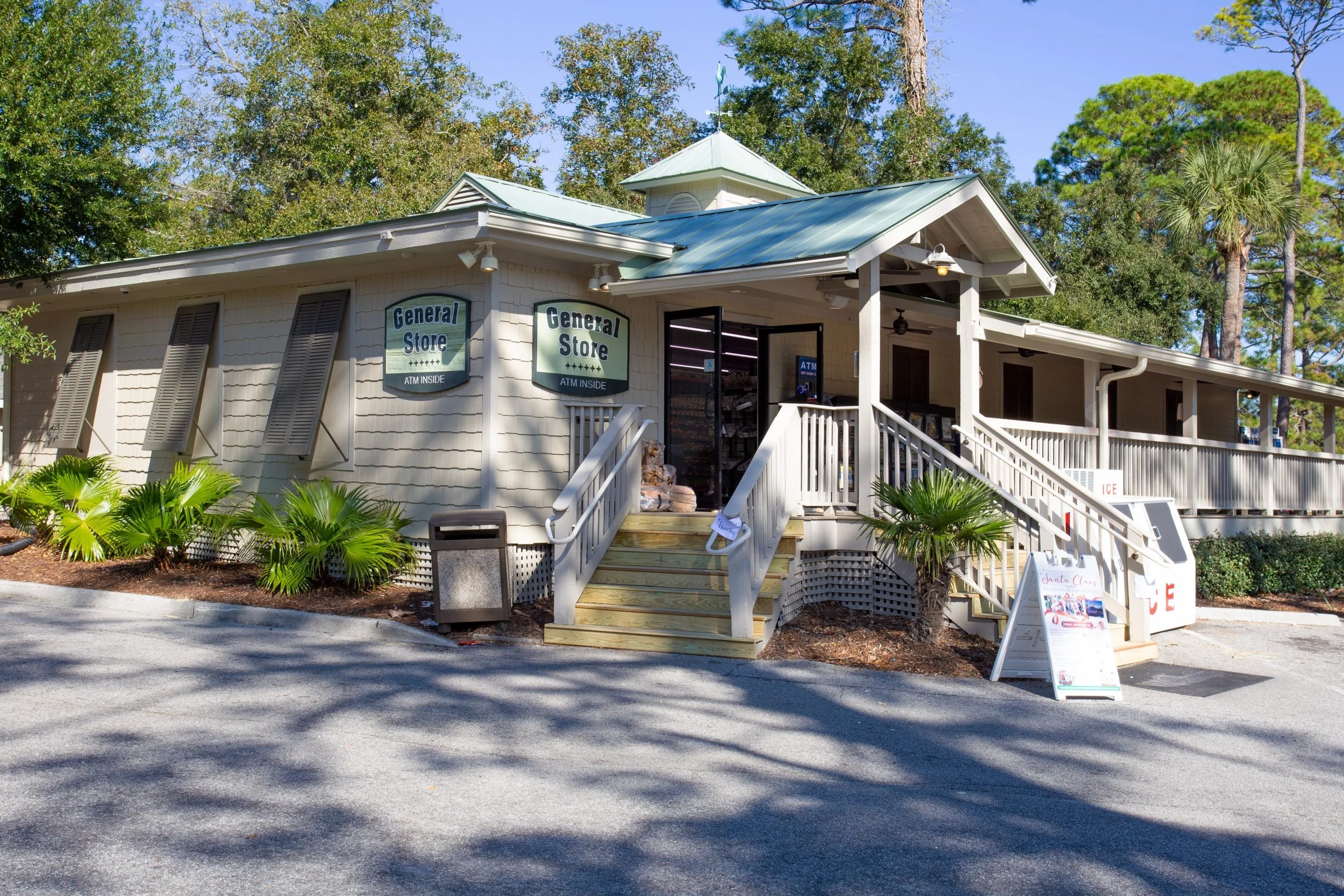 A general store with a green roof and outdoor staircase, surrounded by trees, featuring signs advertising an ATM inside.
