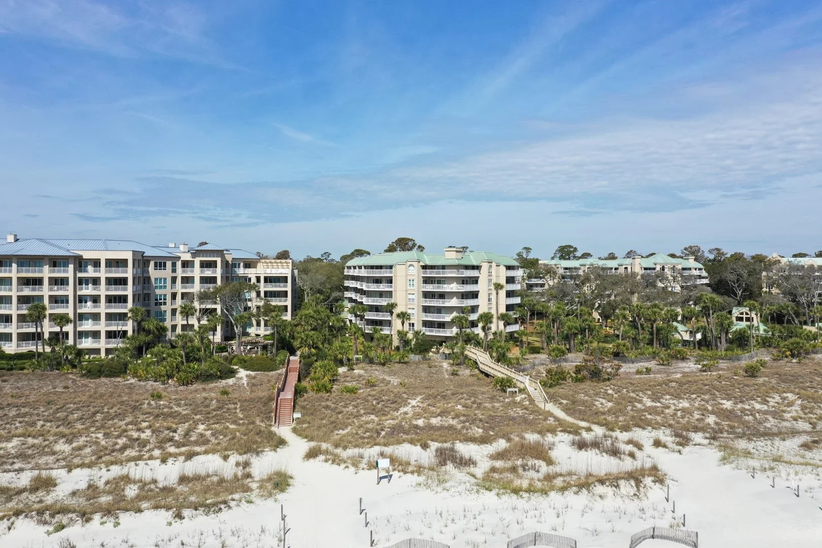 Coastal condominium buildings surrounded by greenery and palm trees, with sandy dunes in the foreground under a blue sky.