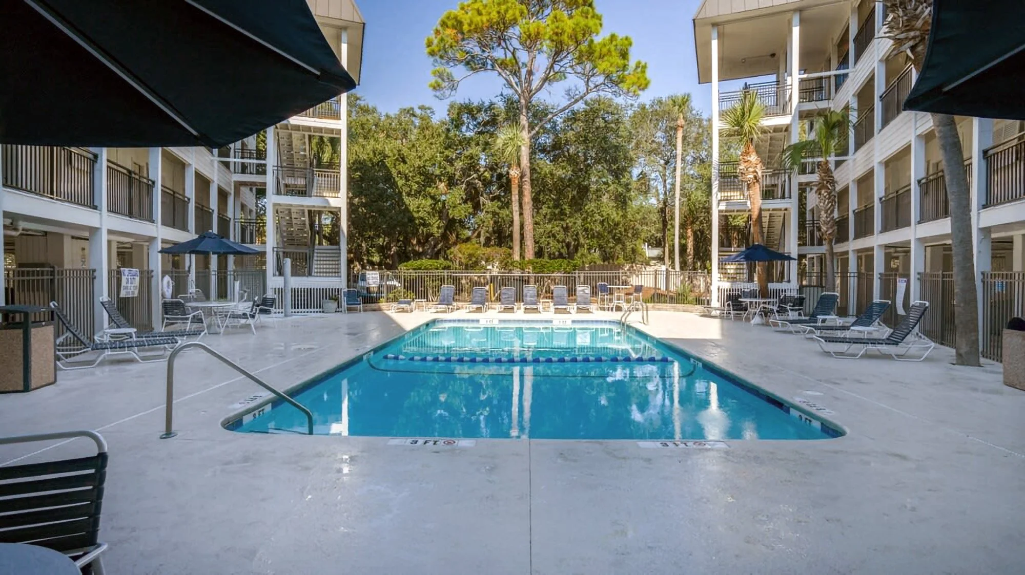 Outdoor swimming pool surrounded by lounge chairs and multi-story buildings, with trees in the background.