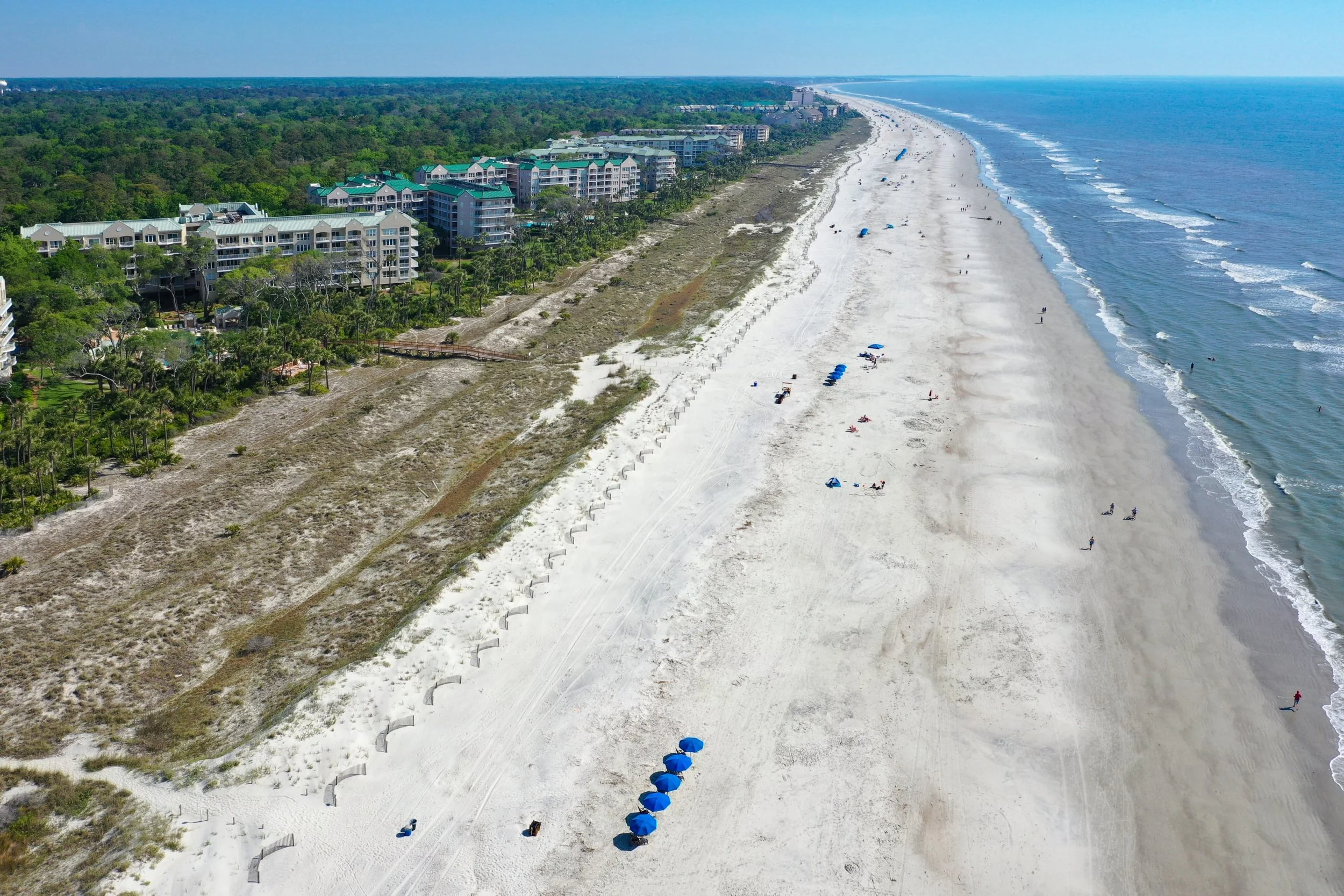 Aerial view of a beach lined with resorts and large buildings, featuring sandy shores, ocean waves, beach umbrellas, and scattered people along the coastline.