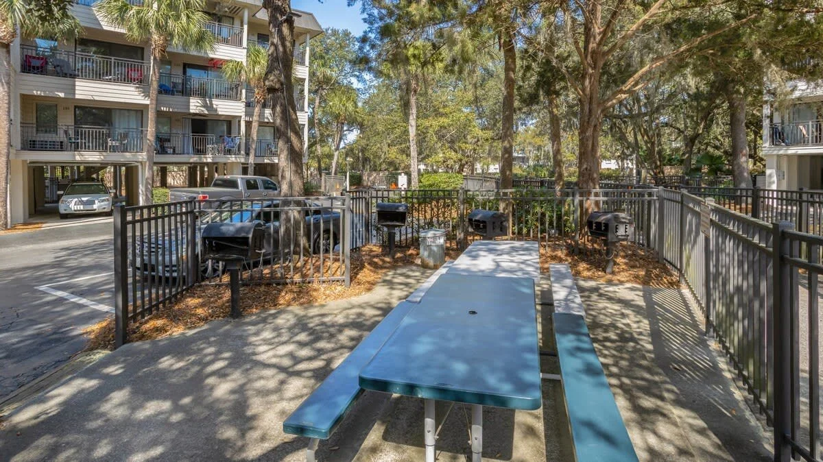 Outdoor picnic area with table, grills, and benches surrounded by trees, near an apartment building and parked cars.
