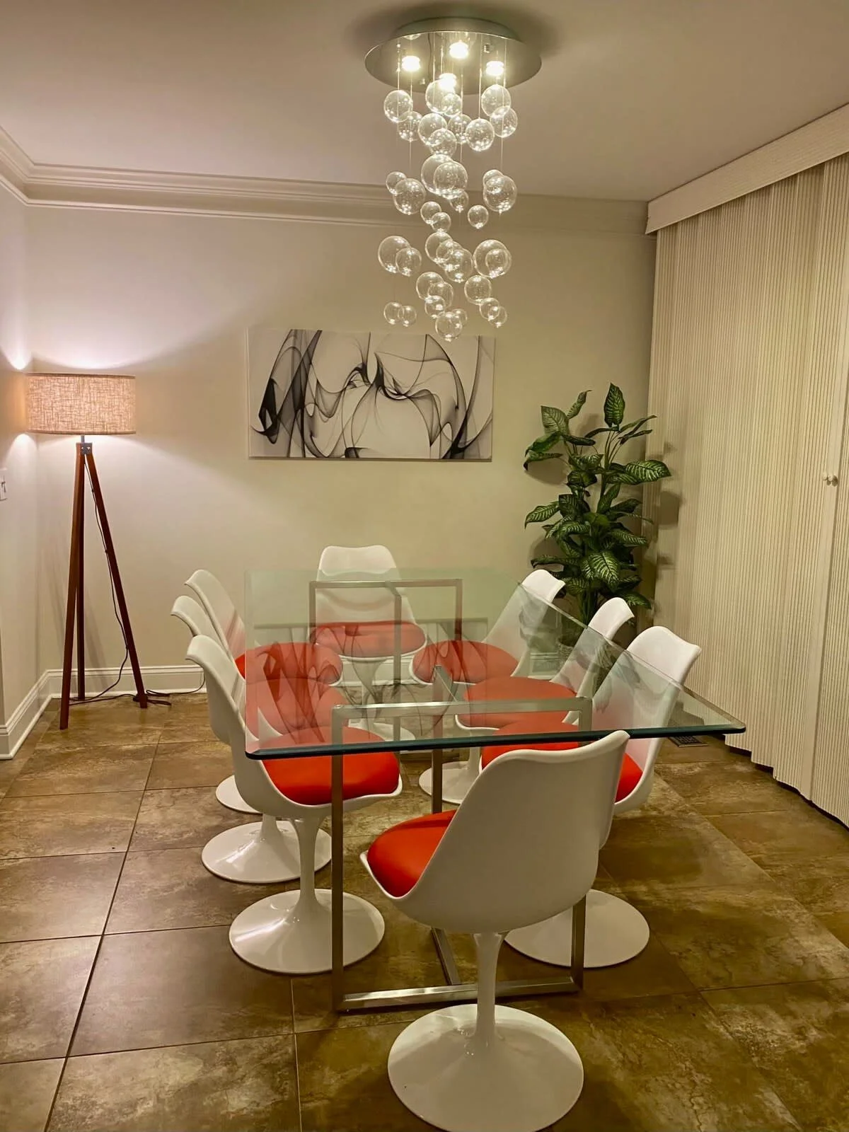Modern dining room with glass table and white chairs with red cushions, artistic chandelier with glass spheres, abstract wall art, floor lamp, and a potted plant.