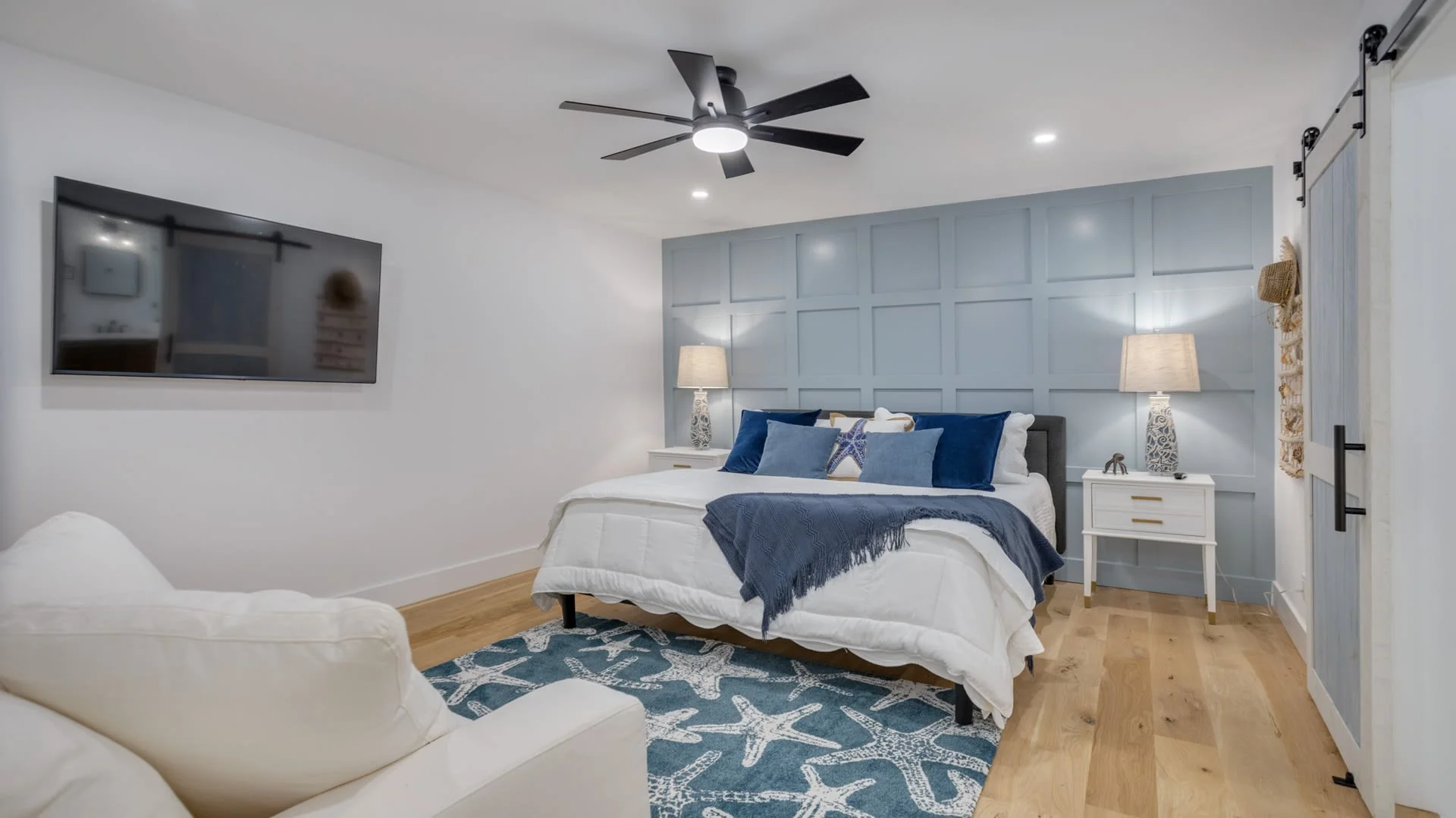 Modern bedroom with blue accent wall, queen bed with blue and white bedding, two bedside tables with lamps, wall-mounted TV, white armchair, wood floor, and nautical-themed rug.