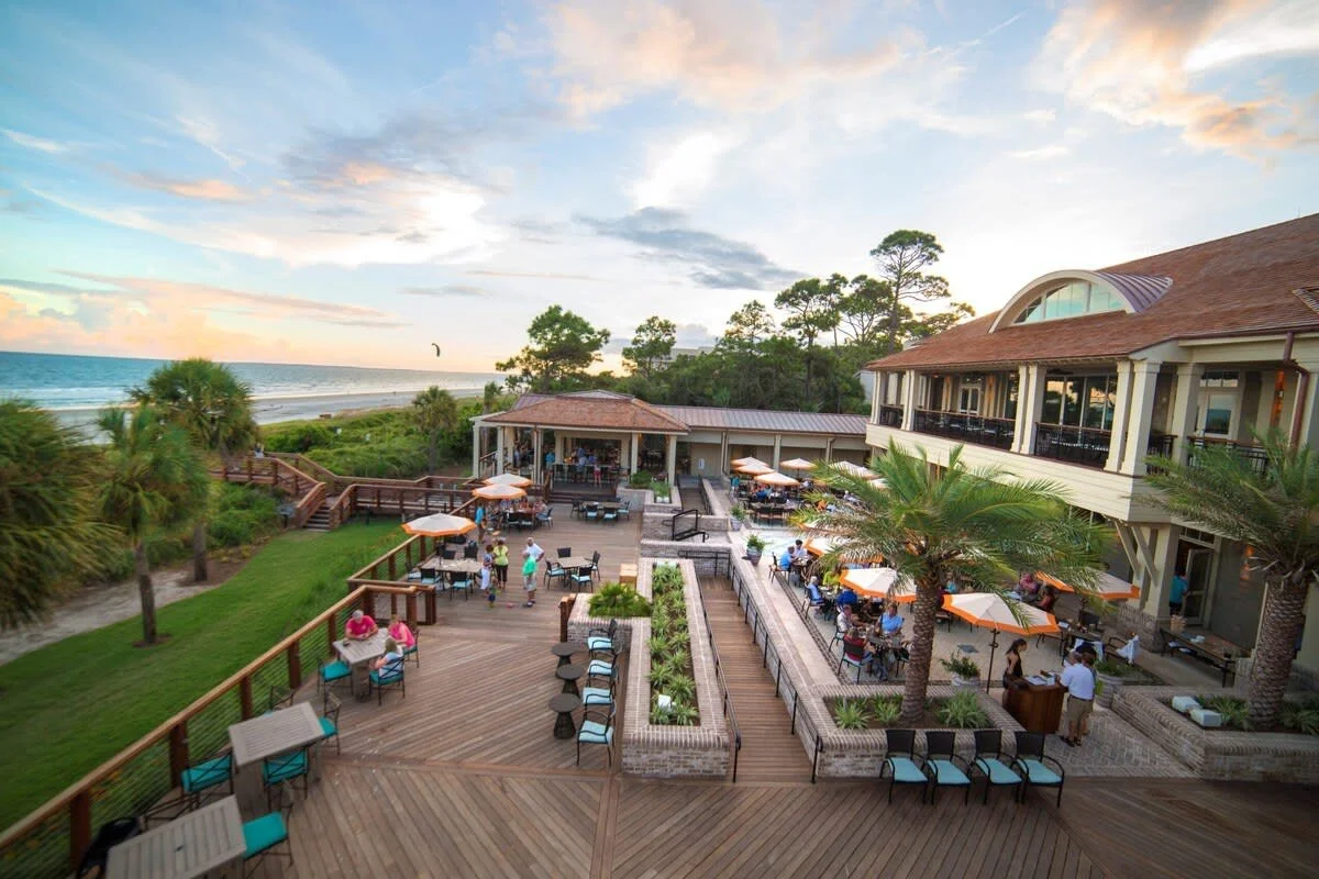 Aerial view of beachfront resort with outdoor dining area, people seated under orange umbrellas, surrounded by lush greenery and palm trees, overlooking the ocean and sandy beach.