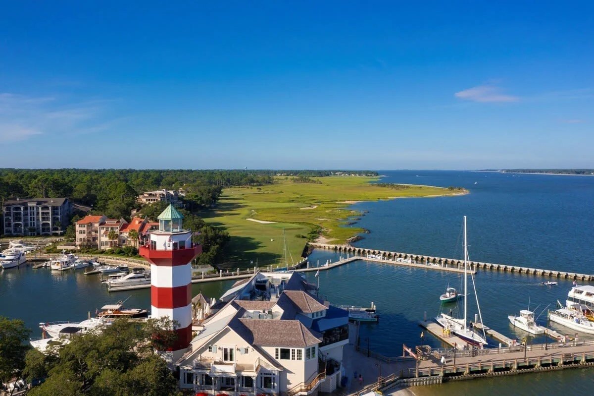 Aerial view of a marina with a red and white lighthouse, boats docked, buildings, and a coastal landscape with greenery and blue water.