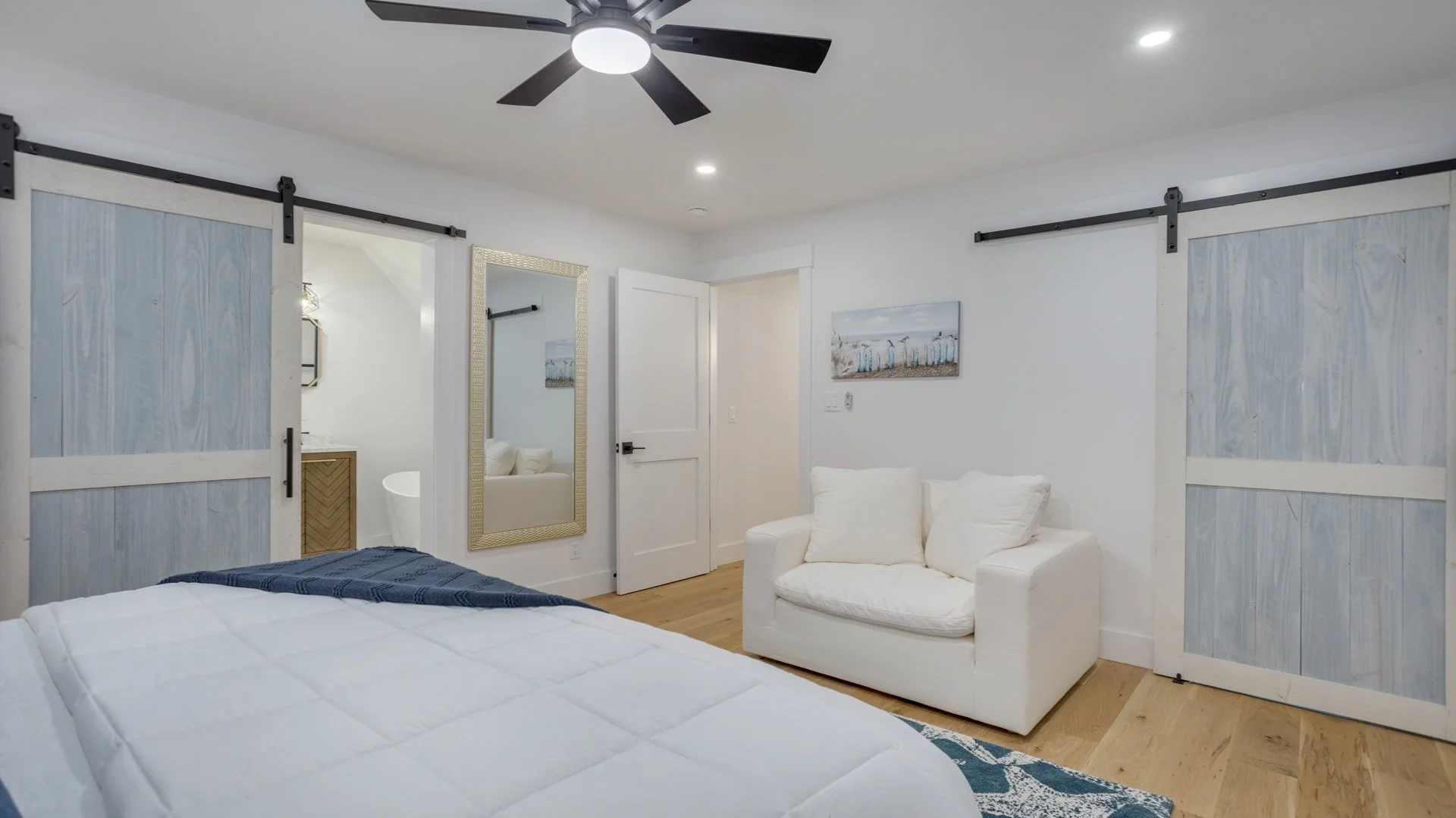 Modern bedroom with a white armchair, barn-style sliding doors, a ceiling fan, a large wall mirror, framed artwork, and a partially visible bed with a white quilt and blue throw blanket.