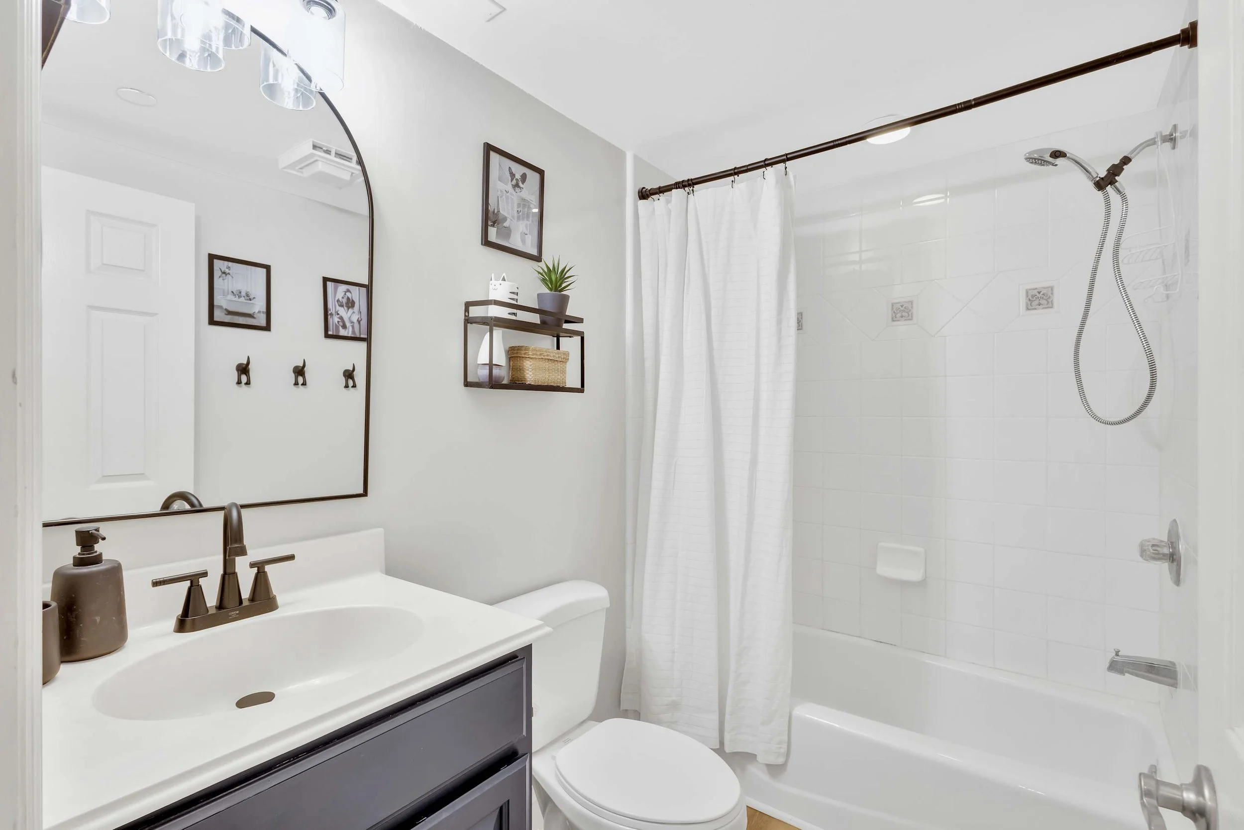 Modern bathroom with white tile bathtub and shower, white curtain, black framed mirror, sink with black faucet, toilet, wall shelves with decor, and framed art.