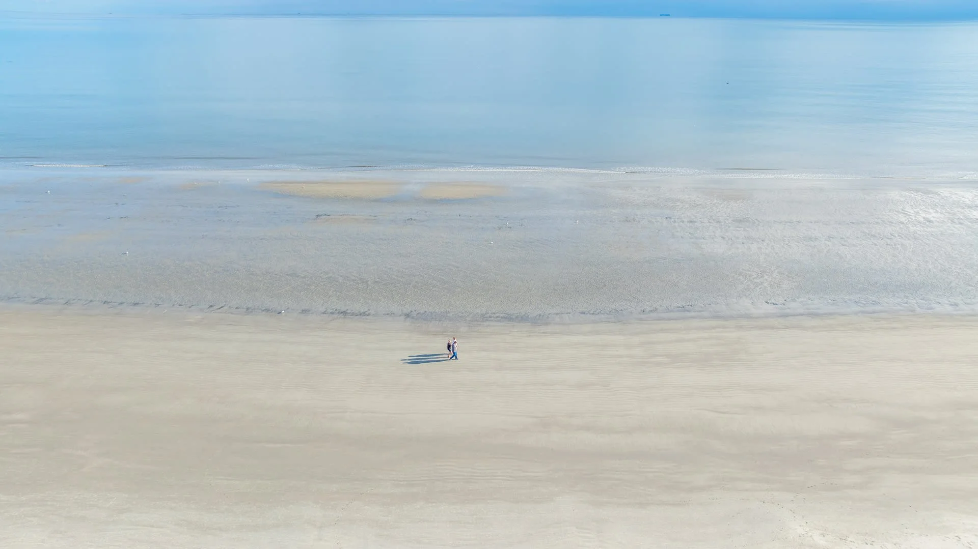 Aerial view of a tranquil beach with two people walking along the sandy shoreline, with calm blue ocean waves in the background.