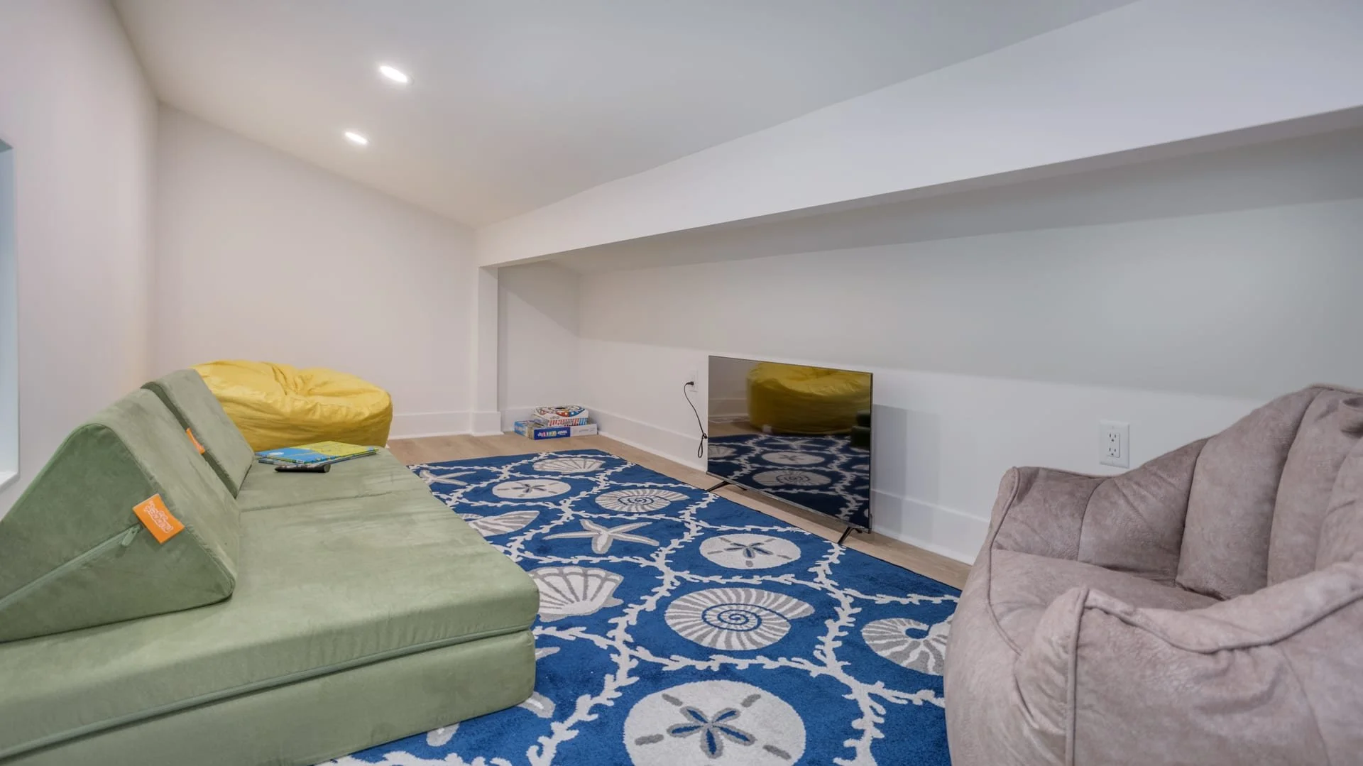 Cozy attic playroom with a green floor couch, yellow bean bag, gray bean bag chair, blue patterned rug, board games, and a flat-screen TV.