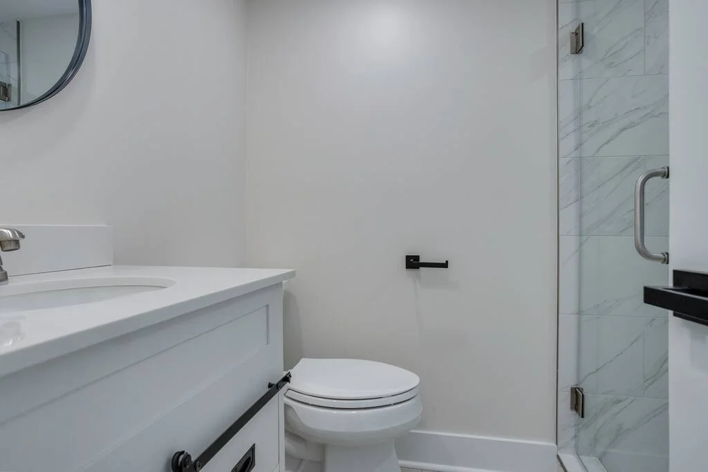 Modern bathroom with white vanity, round mirror, toilet, and glass shower enclosure.