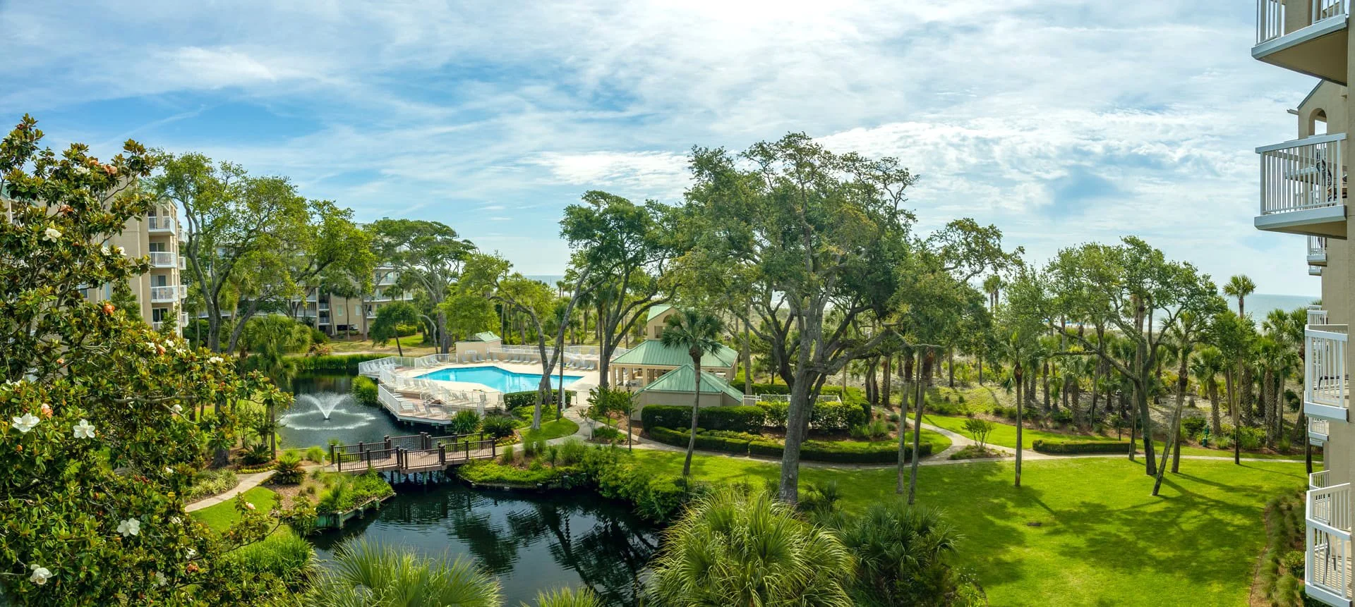 Aerial view of a resort with a swimming pool, surrounded by lush greenery and tall trees. There are beige buildings with balconies in the background, and a small pond with a fountain and a wooden bridge in the foreground.