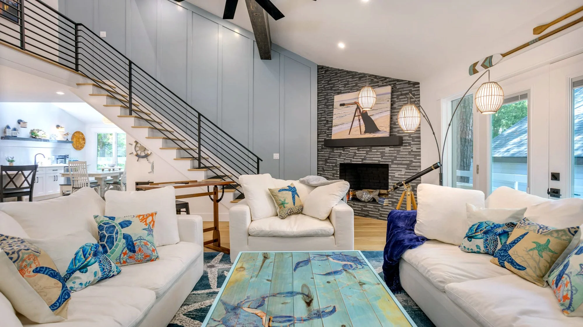 Contemporary living room with white sofas, a colorful wooden coffee table, decorative pillows, a staircase with black railing, and a stone fireplace. The space includes a telescope and coastal-themed decor.