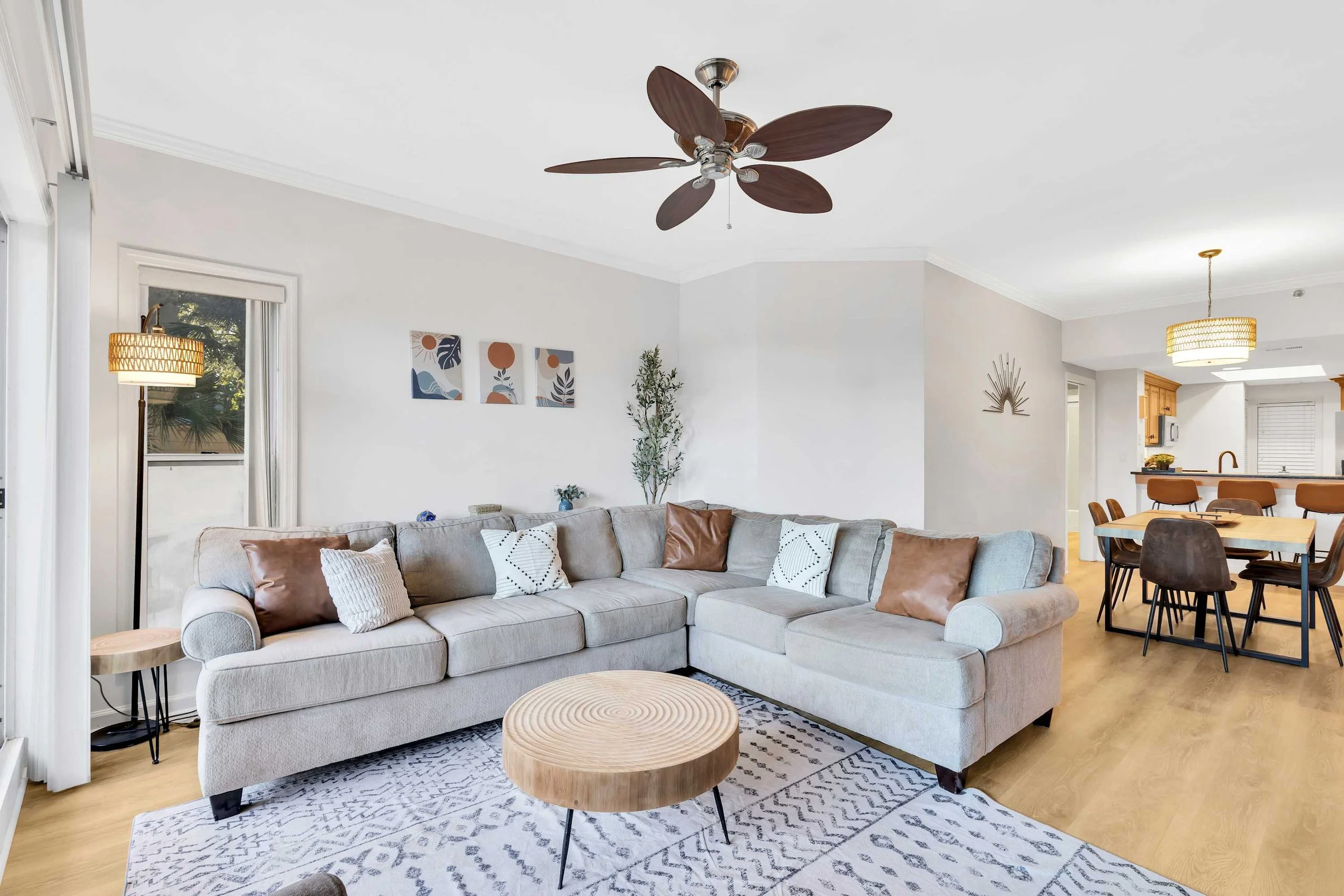 Modern living room with a gray sectional sofa, decorative pillows, a wooden round coffee table, abstract wall art, and a ceiling fan. Adjacent dining area with a table, chairs, and pendant light. Floor lamp by the window, inside a well-lit room with 