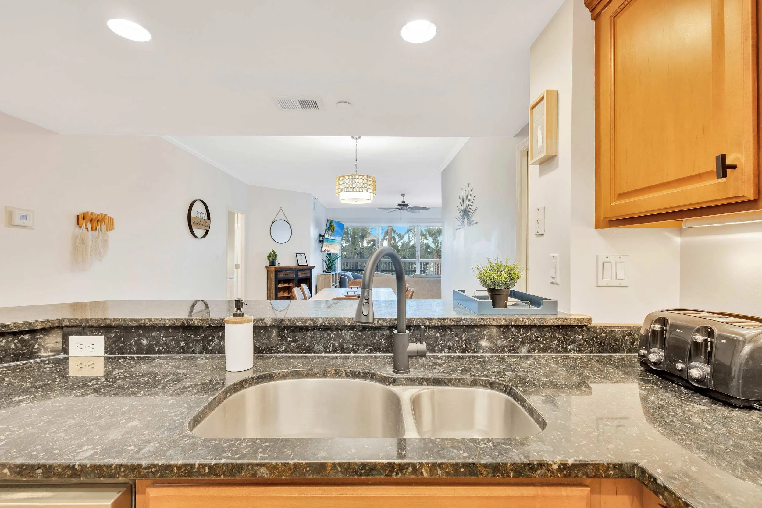 Modern kitchen with granite countertops, stainless steel sink, and a black faucet. A toaster is on the counter next to light wood cabinets. The kitchen opens to a living area with natural light, ceiling fans, and decorative items.