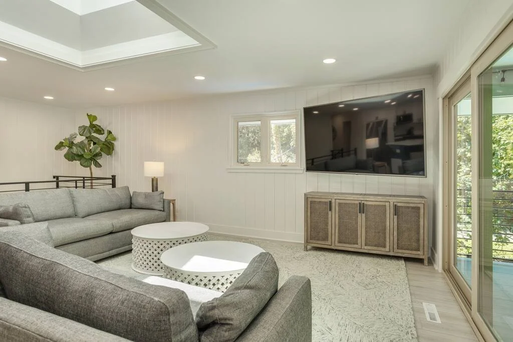 Modern living room with gray sectional sofa, white round coffee tables, large wall-mounted TV, and wooden cabinet, featuring a plant and a window. The space is well-lit with natural light from adjacent sliding glass doors.