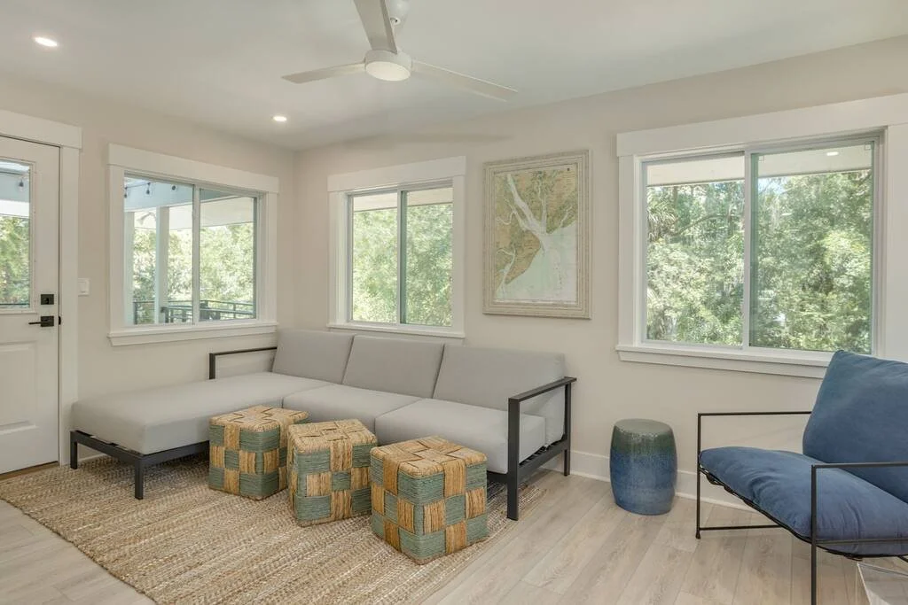 Modern living room with gray sectional sofa, woven ottomans, blue armchair, wall map, large windows, and light wood flooring.