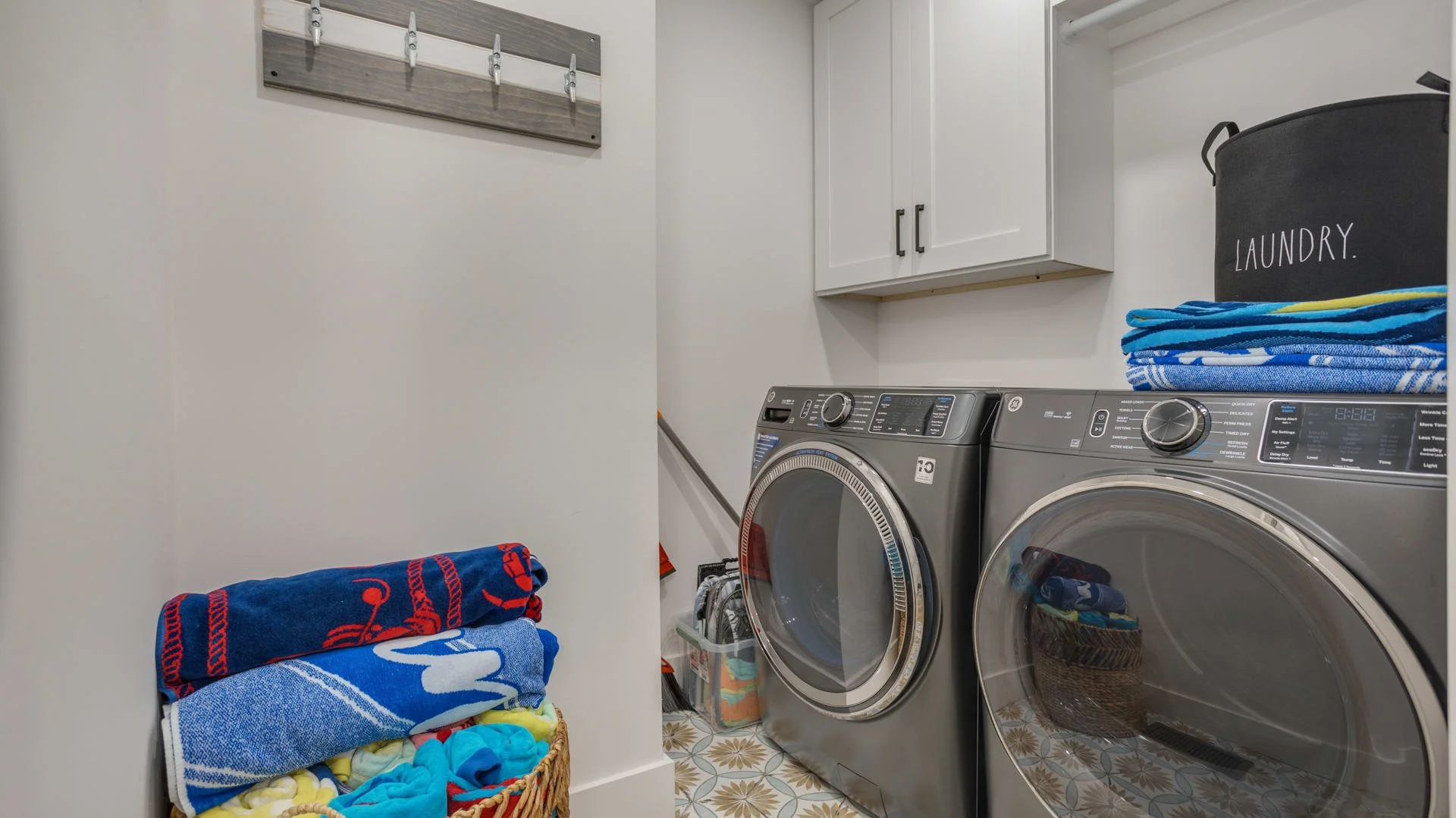 Laundry room with washer and dryer, folded towels, wall hooks, and storage bin labeled 'LAUNDRY'.