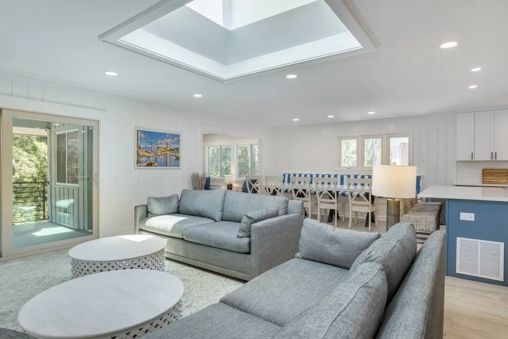 Modern living room with gray sofas, round coffee tables, large skylight, and a dining area with blue-striped chairs.