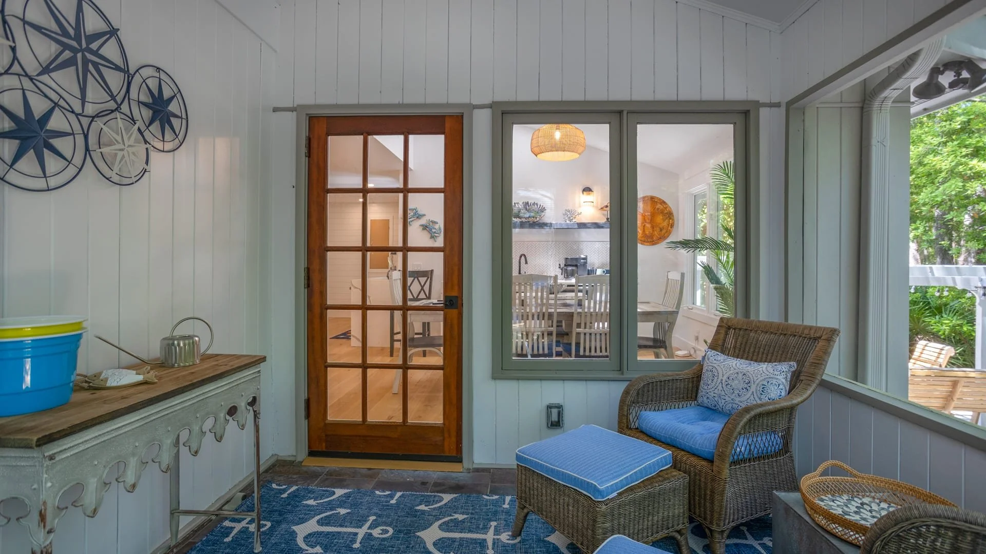 Cozy sunroom with wicker furniture, nautical-themed decor, and a wooden door leading to a bright dining area inside. Walls are white paneled, featuring star-shaped wall art.