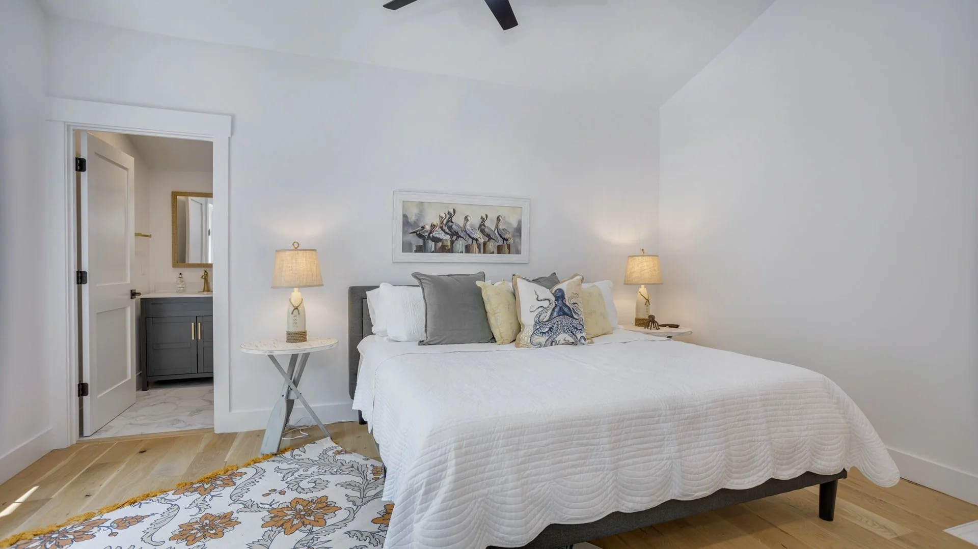 A bedroom with a neatly made bed with white and gray pillows, two bedside tables with lamps, a piece of wall art depicting birds, a wooden floor with a patterned rug, and an open door leading to a bathroom.