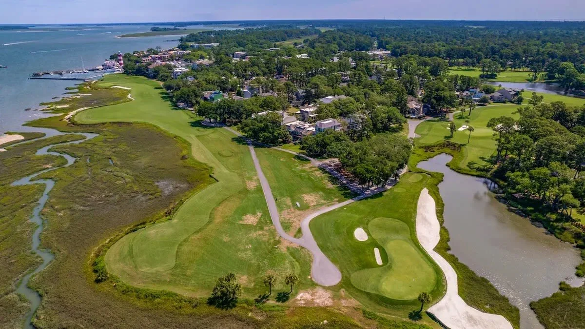 Aerial view of a coastal golf course with greens, sand traps, and water hazards surrounded by trees and residential buildings, adjacent to an ocean or large body of water.