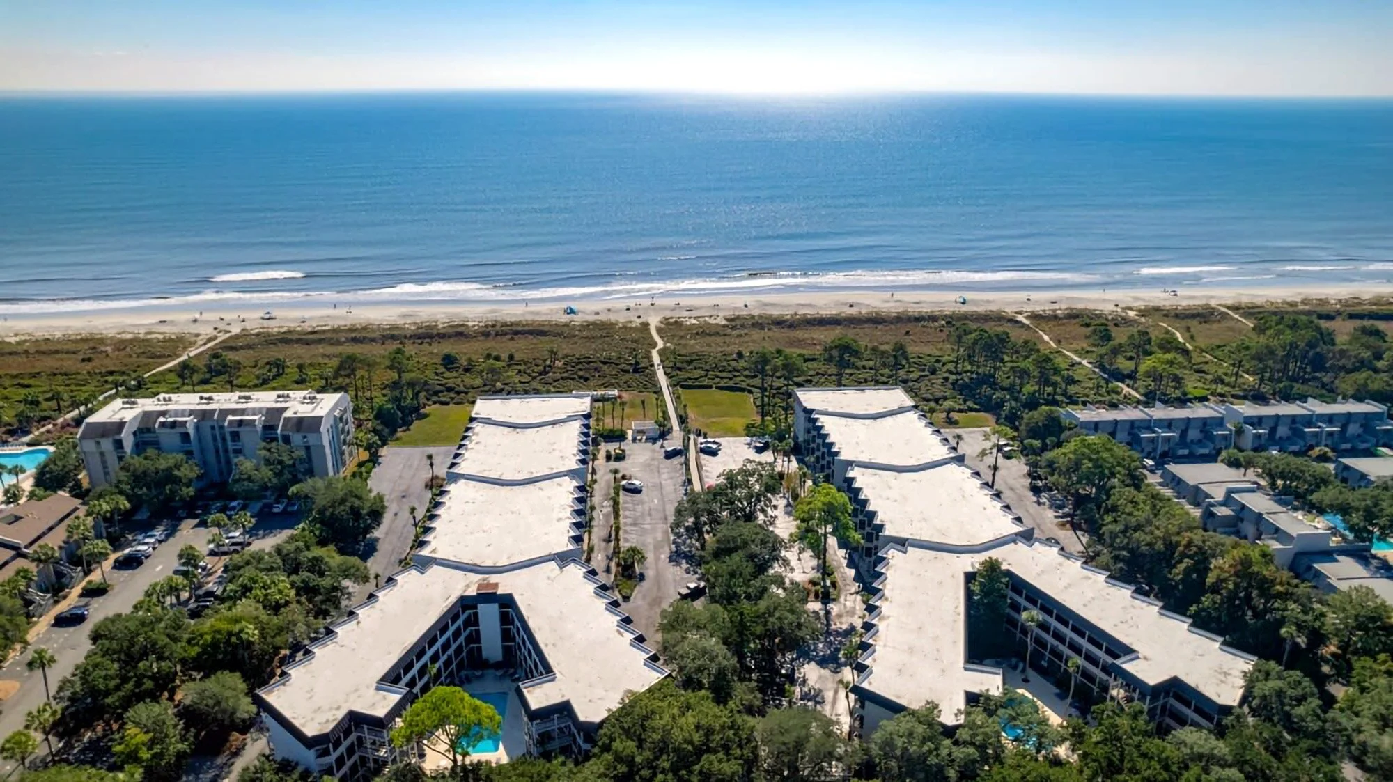 Aerial view of beachfront resort with buildings, palm trees, and a coastline leading to a sandy beach and the ocean.