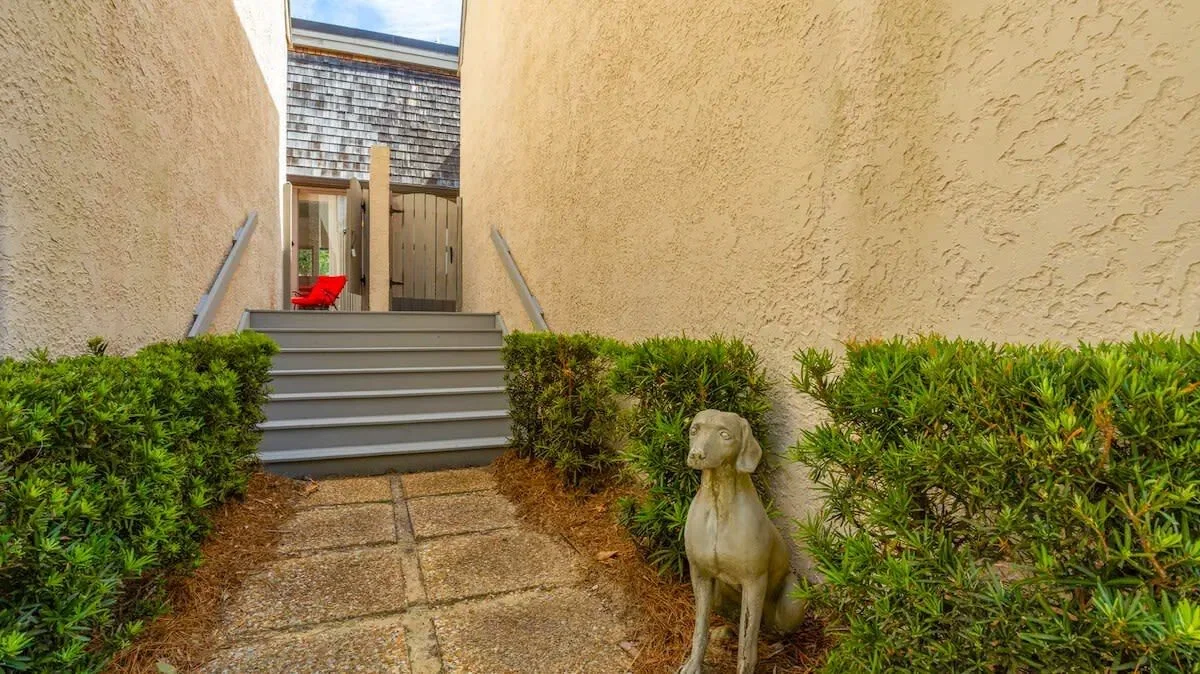 Pathway between stucco walls with greenery, leading to steps and a wooden gate; a dog statue is on the right.