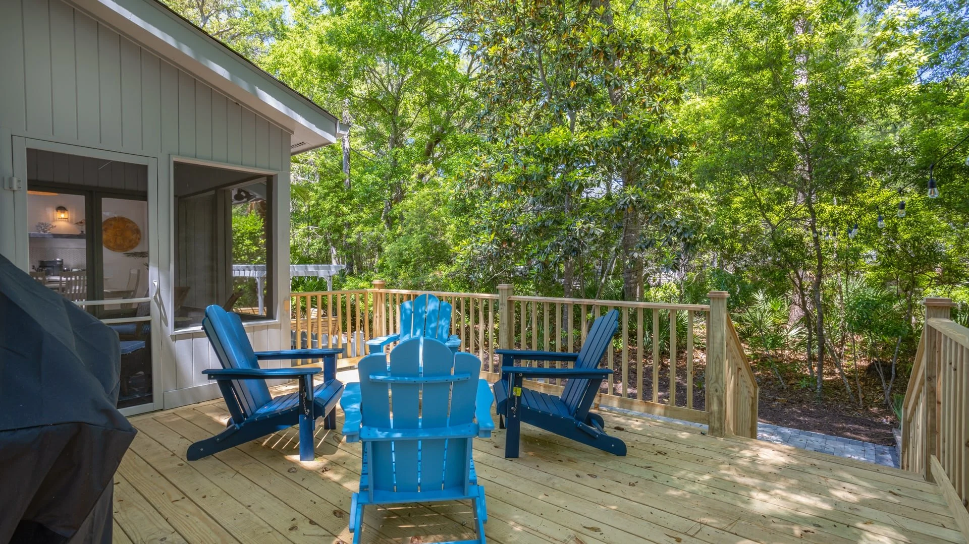 Outdoor deck with blue Adirondack chairs, surrounded by trees, featuring a wooden railing and strings of lights.