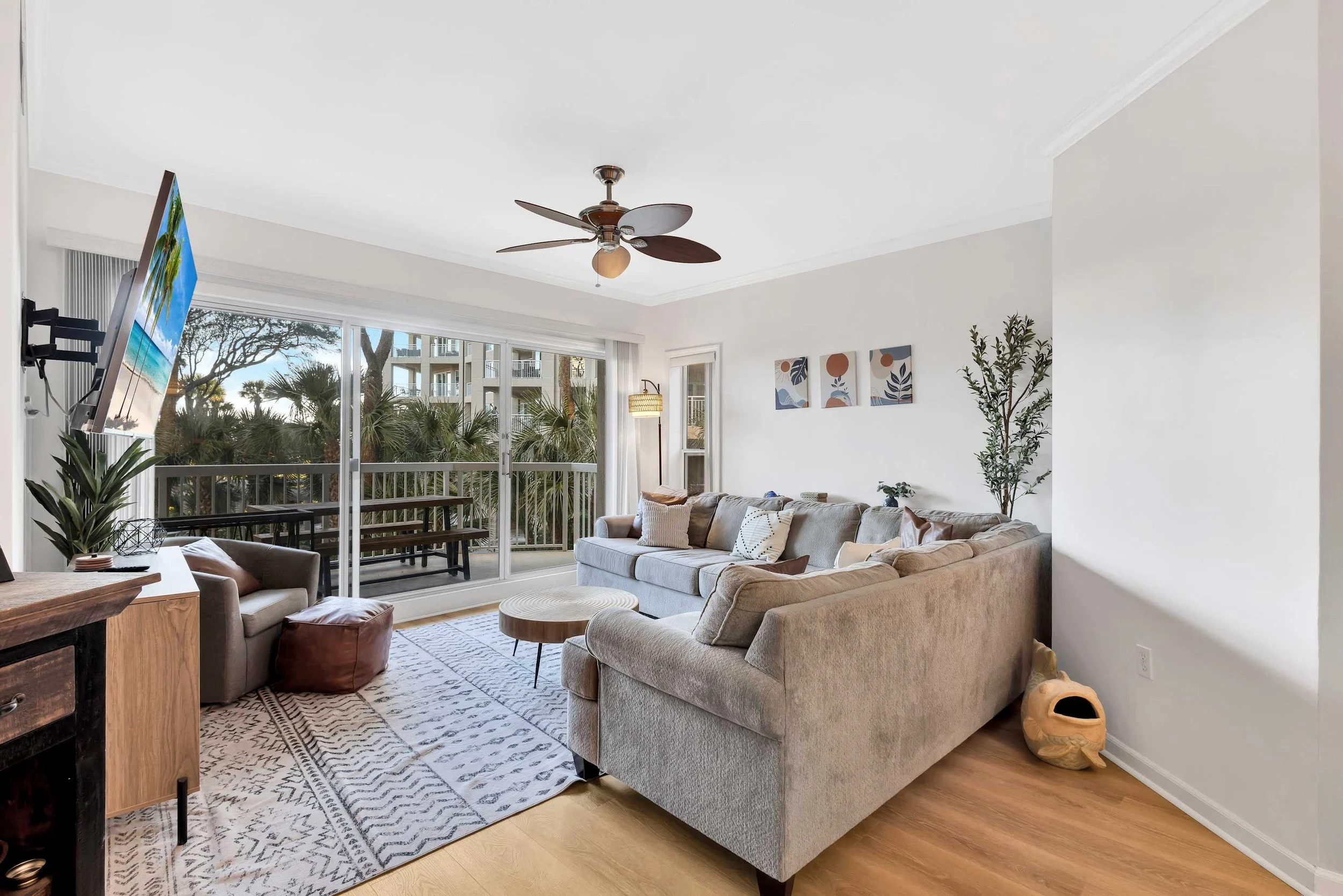 Modern living room with large gray sectional sofa, patterned rug, wall art, and ceiling fan. Large sliding glass doors open to a balcony with outdoor furniture and palm tree views.