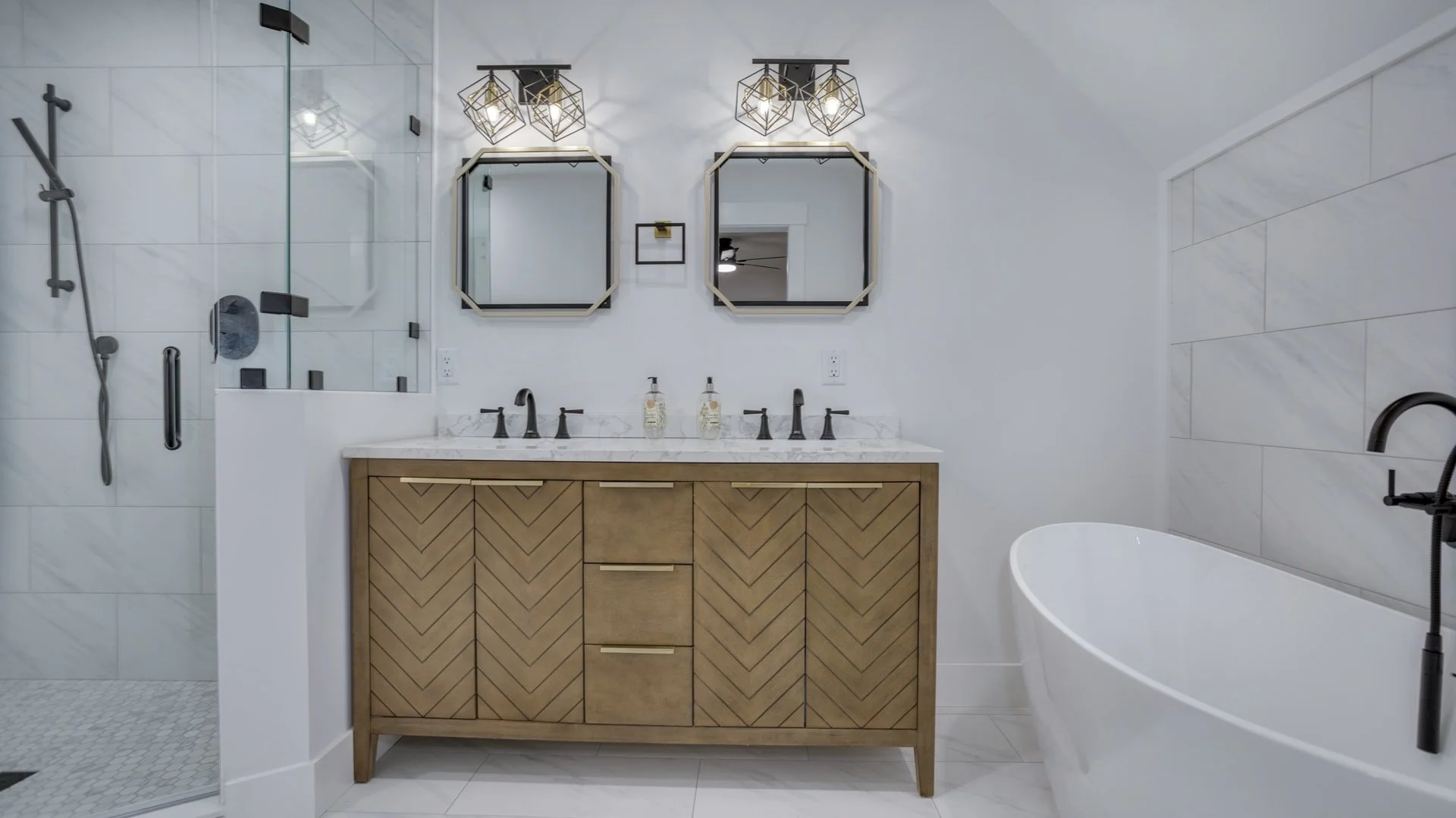 Modern bathroom with double vanity, marble countertop, chevron-patterned cabinet, two geometric mirrors, wall-mounted light fixtures, glass-enclosed shower, and standalone white bathtub.