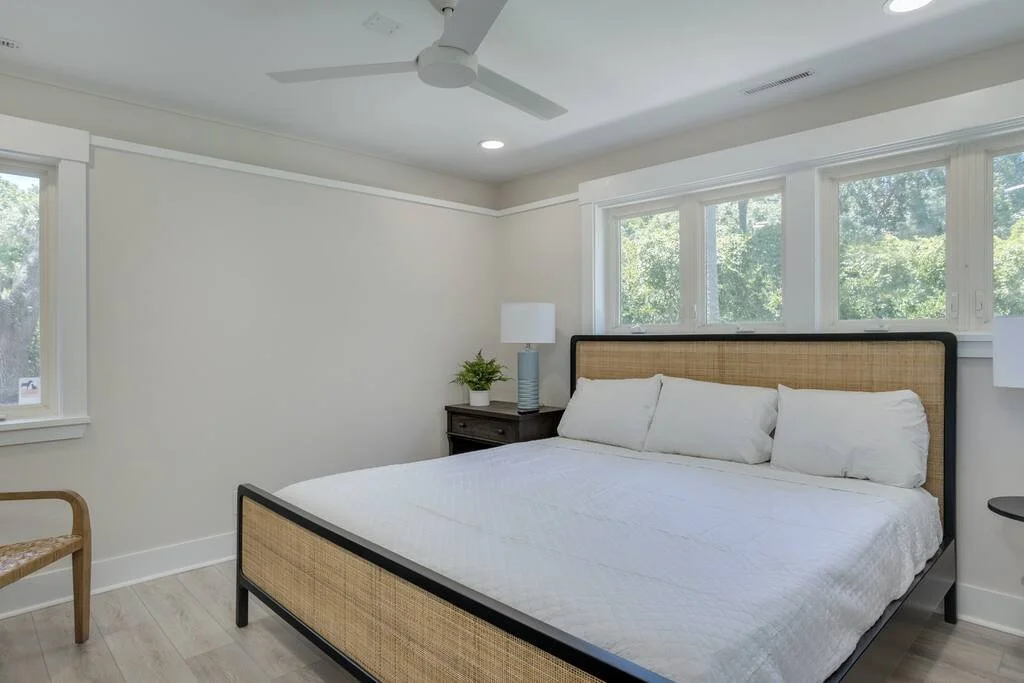 Bright bedroom with white walls, large bed, wicker headboard, wooden side chair, and a nightstand with a lamp and plant, featuring three windows and a ceiling fan.