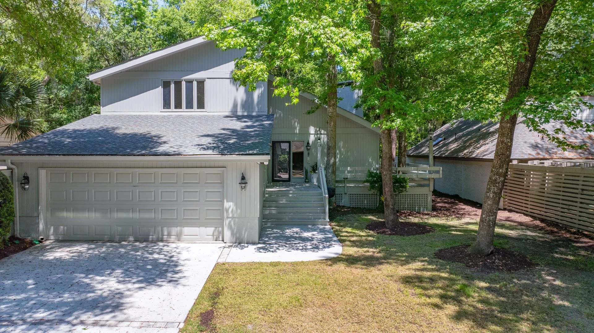 A modern two-story house with a light gray exterior and a dark shingled roof. The front features a two-car garage and a set of steps leading to the front door. The property is surrounded by lush greenery and trees, providing shade.