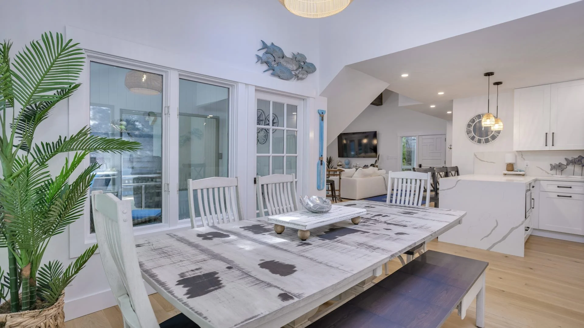 Modern dining room with rustic table, white chairs, potted plant, decorative wall art, open kitchen, and living area.
