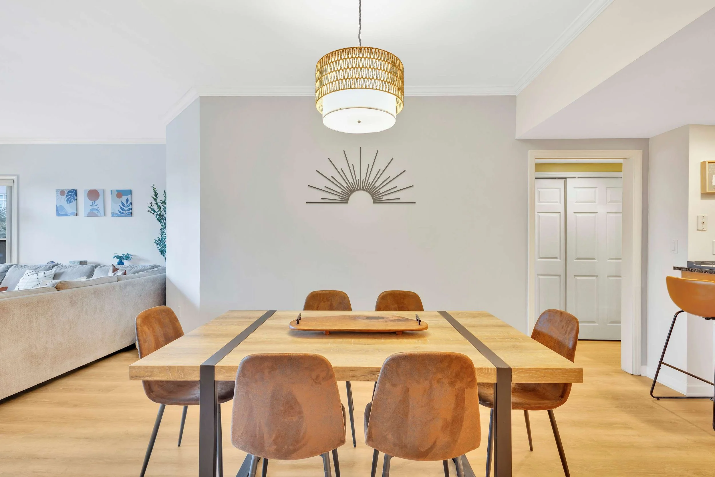 Open dining area with wooden table and six brown chairs, wicker pendant light above, and abstract metal wall art. Adjacent living room with beige sectional sofa, decorative plants, and minimalist art on the wall.