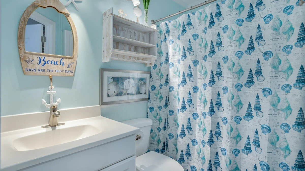 Coastal-themed bathroom with beach decor, featuring a mirror with the phrase 'Beach Days Are the Best Days,' a blue and white shower curtain with seashell patterns, a white sink with an anchor-shaped faucet, and light blue walls.