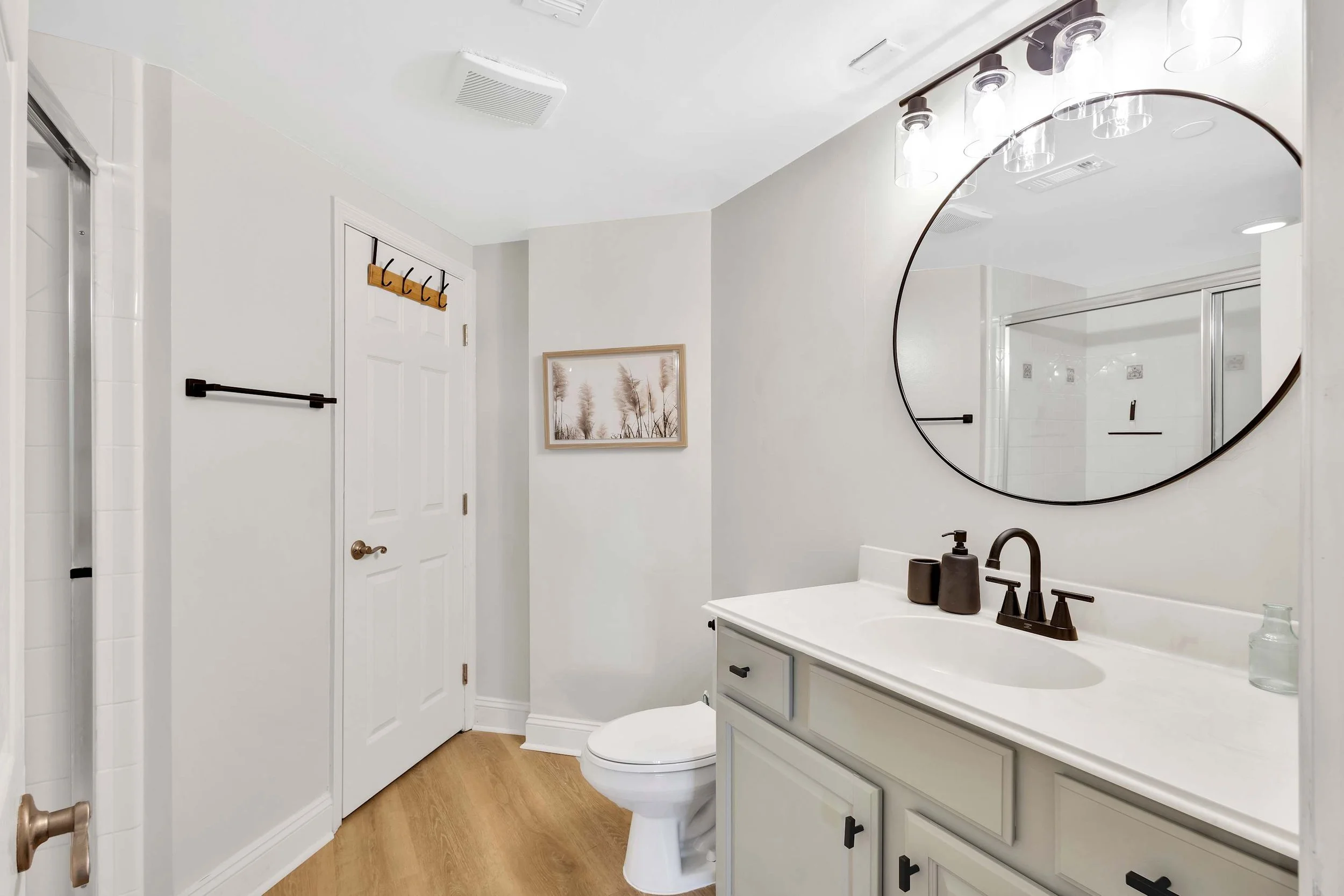 Modern bathroom with large round mirror, white vanity, black fixtures, shower, and framed artwork on wall.