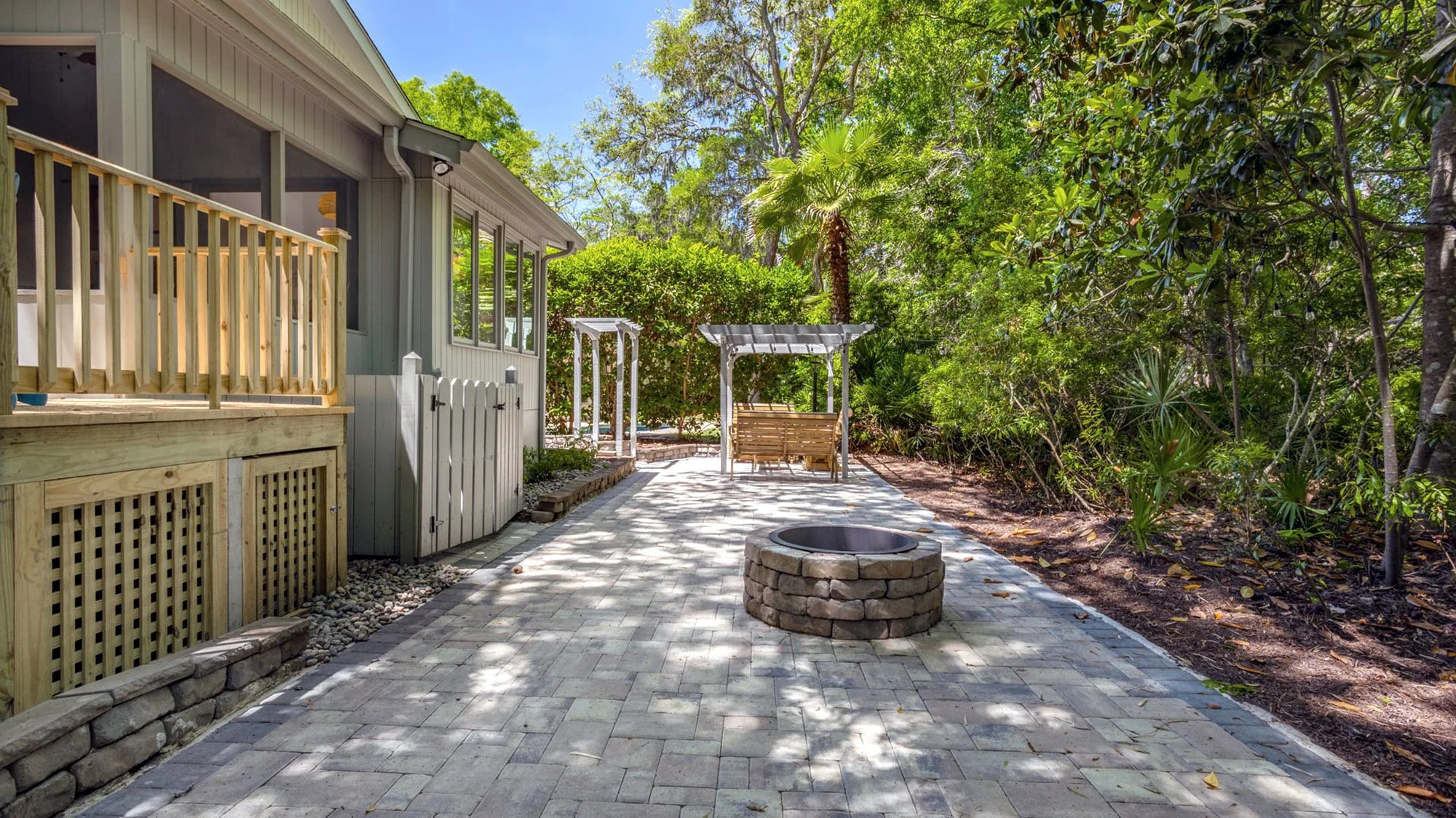Backyard patio area with a brick fire pit, wooden deck with railing, pergola with a swing, and surrounding lush green trees.