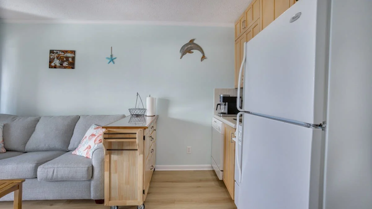 Cozy living room and kitchen area with light blue walls, a gray couch, decorative pillows, wooden furniture, and appliances including a white refrigerator and coffee maker.